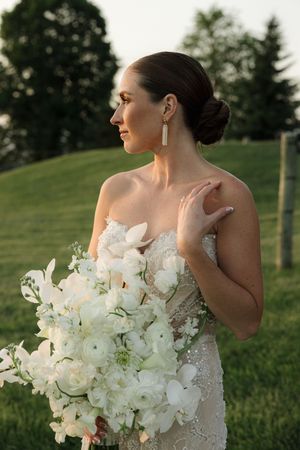 A bride in a strapless lace gown holding an elegant white bouquet of orchids and roses in a garden setting at sunset.