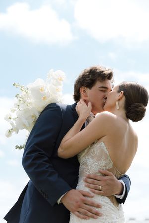 Newlywed couple shares romantic kiss with white bouquet against blue sky background.