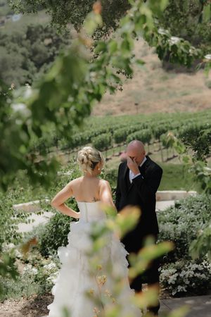 Two figures in wedding attire share an emotional first look moment in a scenic vineyard setting.