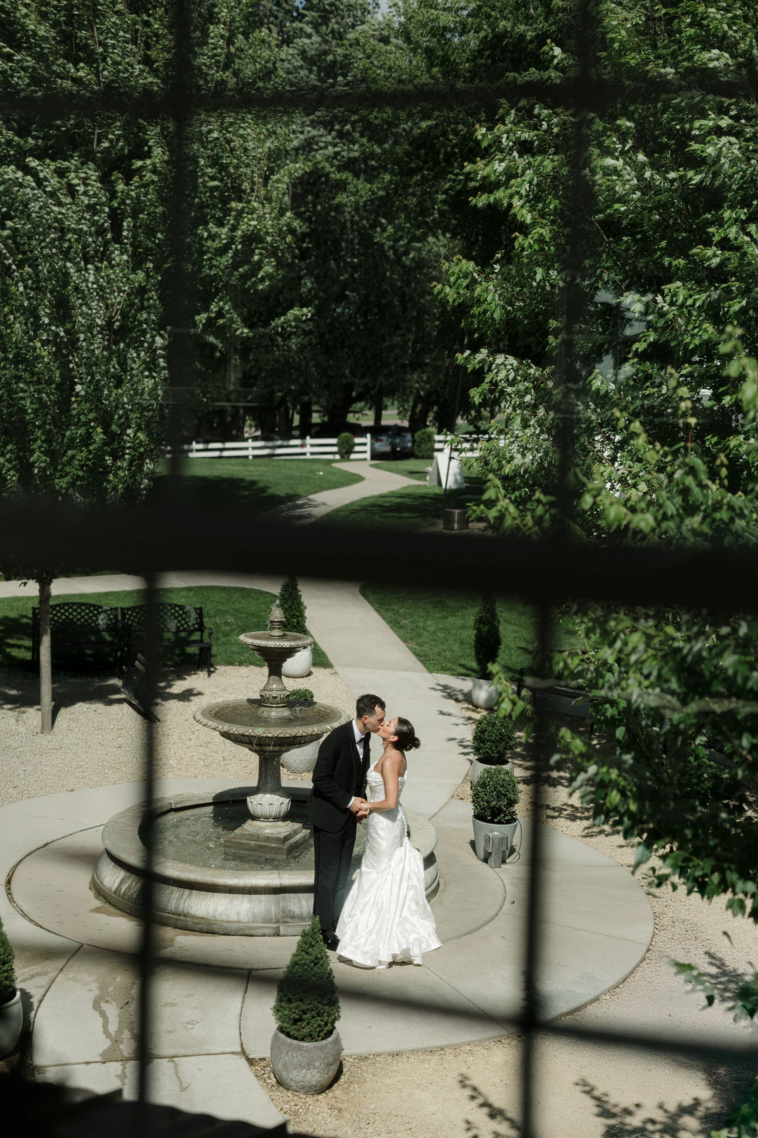 A couple shares an intimate moment in a manicured garden courtyard with stone pavers and elegant fountain features.