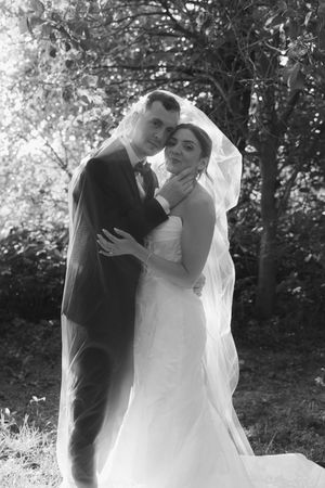 A tender black and white wedding portrait of a couple embracing outdoors with a flowing veil in a natural setting.
