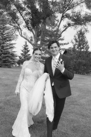 A black and white wedding photo showing a couple in formal attire standing outdoors under a large tree.