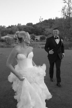 A black and white wedding photo showing a playful moment between bride and groom on a grassy field.