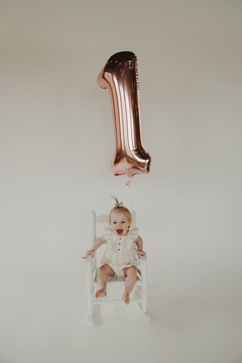 Baby in white outfit sits in rocking chair with rose gold number one balloon floating above for first birthday photo.
