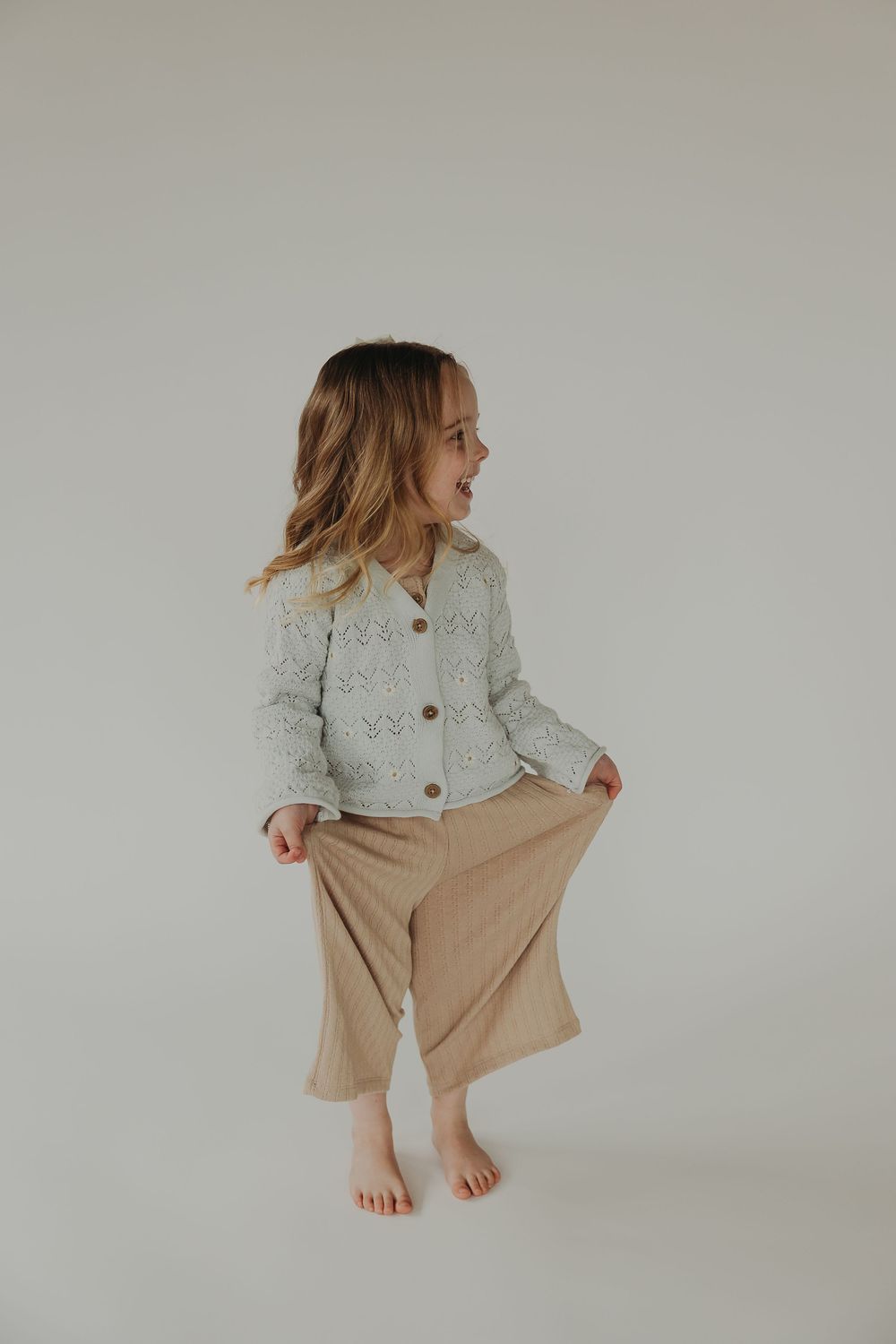 Child wearing mint green button-up cardigan and beige shorts poses playfully against white backdrop in lifestyle shoot.