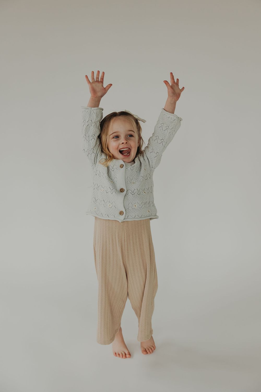 A joyful child wearing a gray cardigan and beige pants jumps with arms raised against a neutral background.
