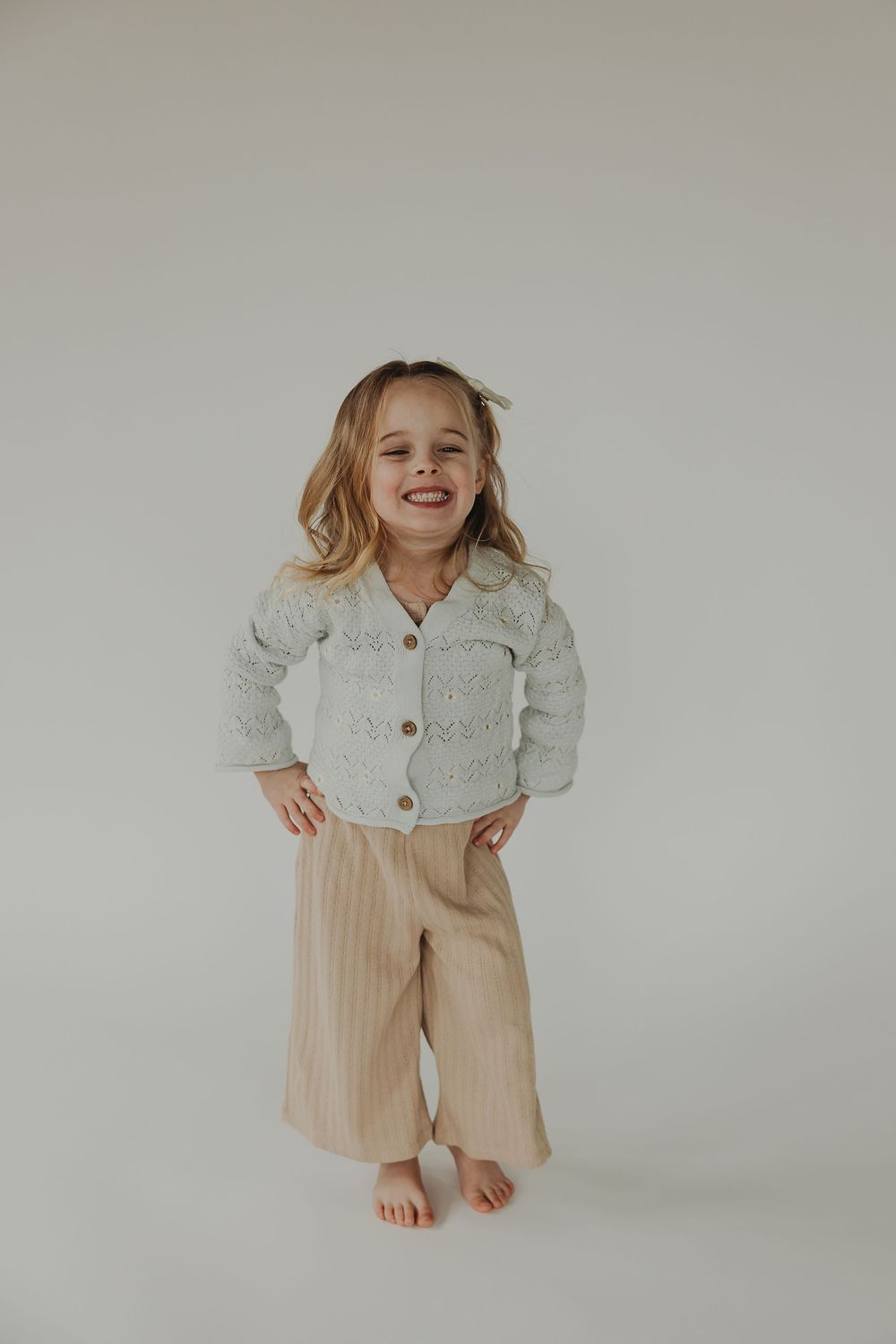A young child in a white knit cardigan and beige striped shorts poses playfully against a light gray backdrop.