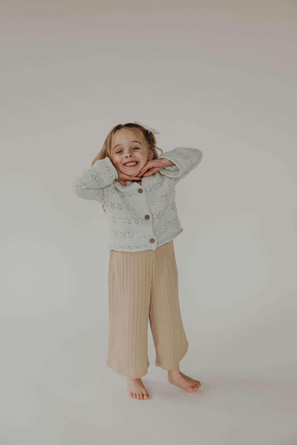 Child wearing a white knit cardigan and beige ribbed pants stands barefoot against light backdrop in playful pose.