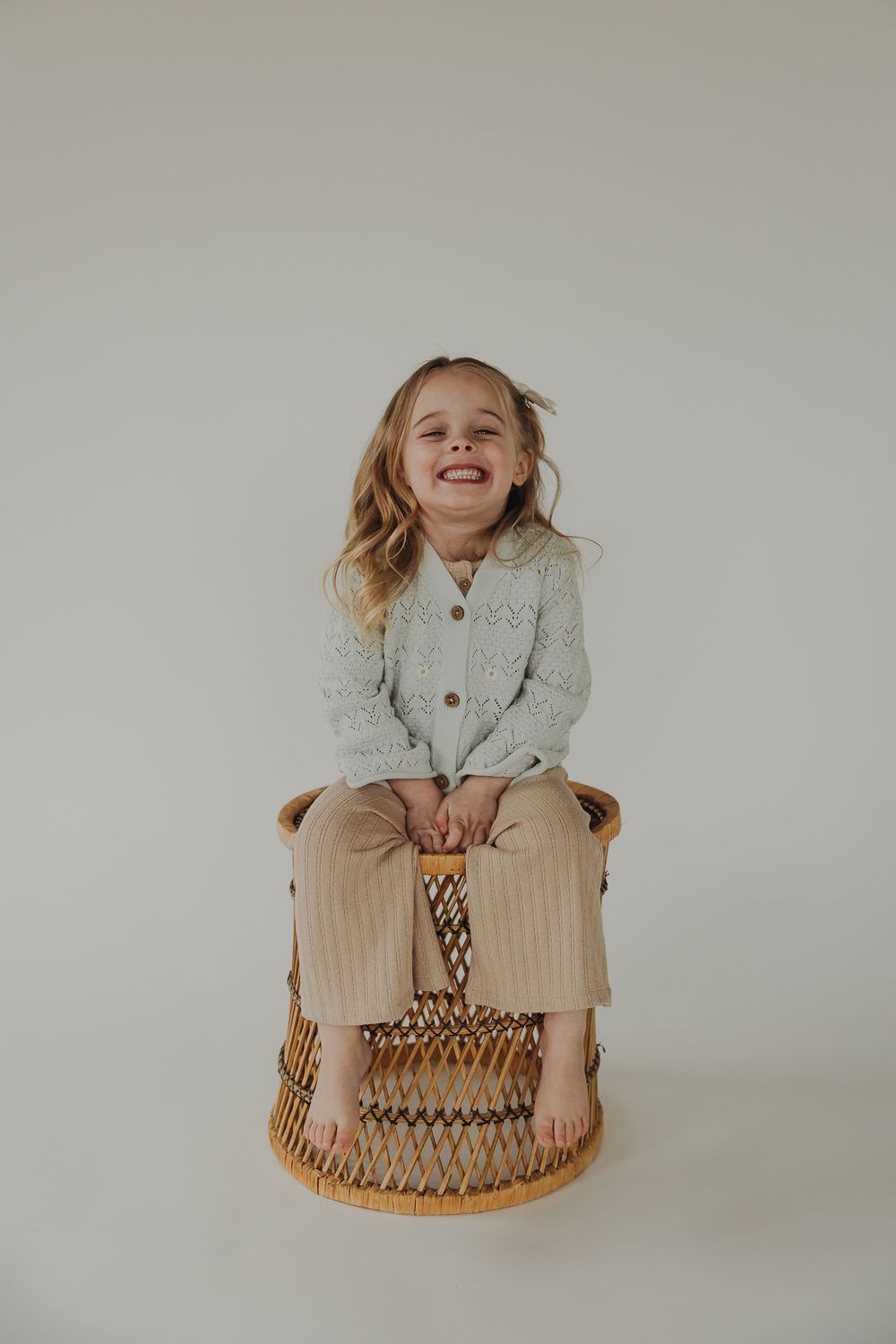 A joyful moment captured on a woven wicker stool against a neutral background, wearing a cozy gray cardigan and khaki pants.