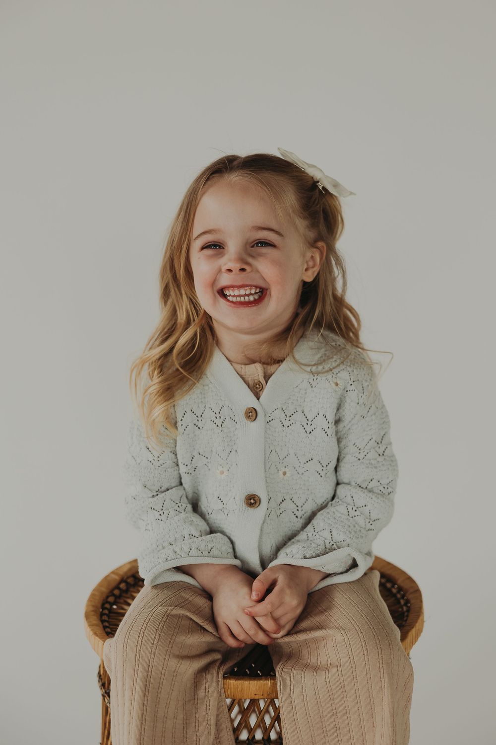 A cheerful young blonde child wearing a light blue cardigan sweater and tan pants sits on a wicker chair against a gray background.