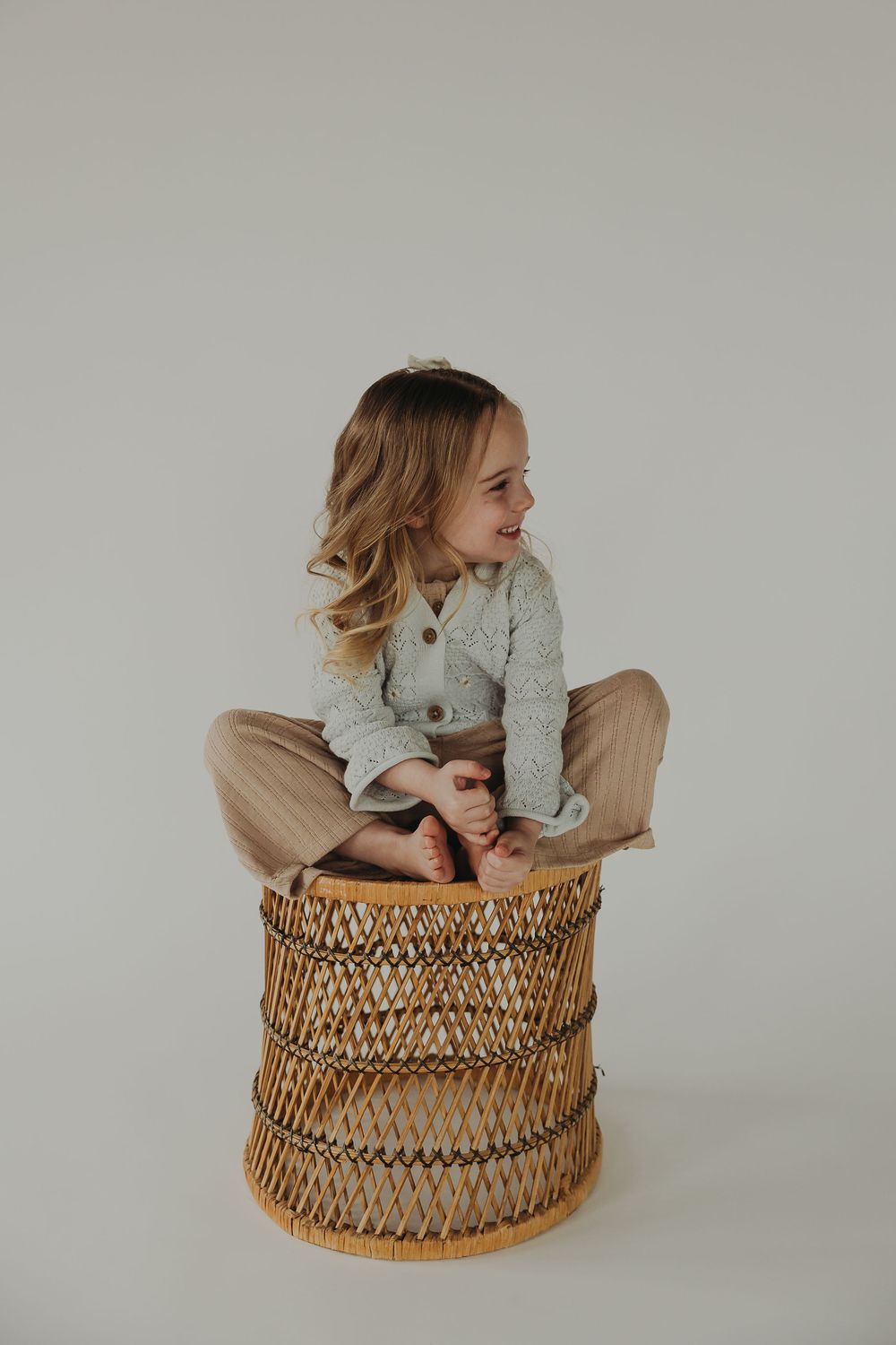 A child in a gray sweater sits cross-legged on a bohemian-style woven rattan stool against a neutral background.