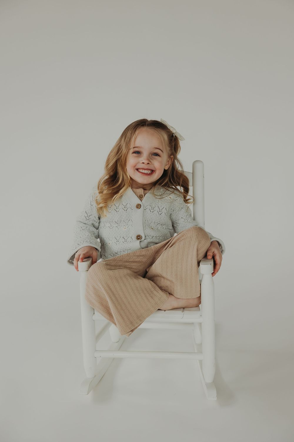 A child in a cozy gray cardigan and beige pants sits and smiles while sitting against a white background.