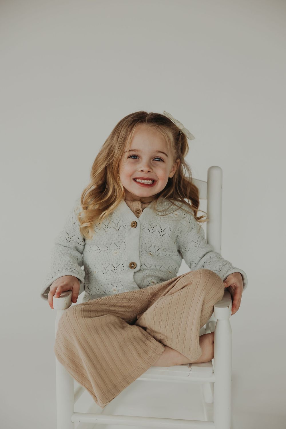 A child in a gray cardigan sweater and beige pants sits in a white wooden chair against a neutral background.