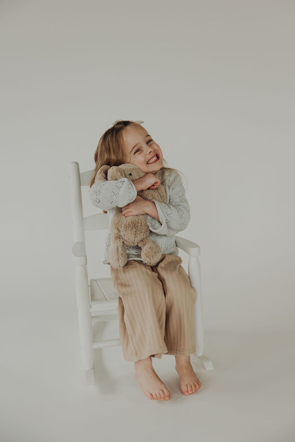 A child in beige pants and gray sweater sits barefoot on a white chair with a playful expression against neutral backdrop.