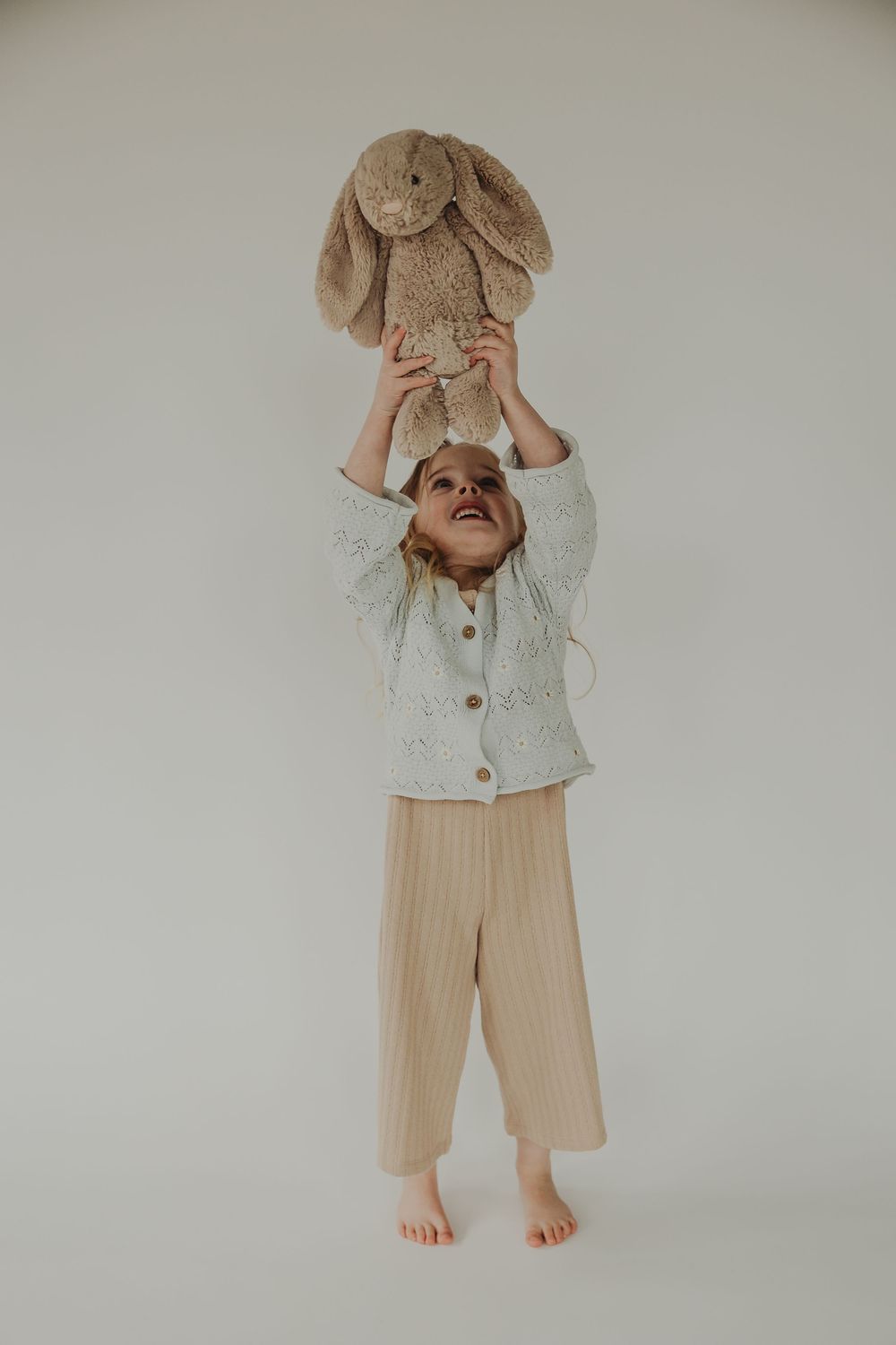 A child in a beige linen outfit is shown from neck down playing peek-a-boo with a soft brown stuffed animal.
