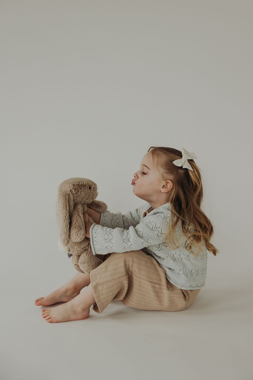 A young child in vintage-style clothing holds up a plush bunny toy while sitting against a neutral backdrop.