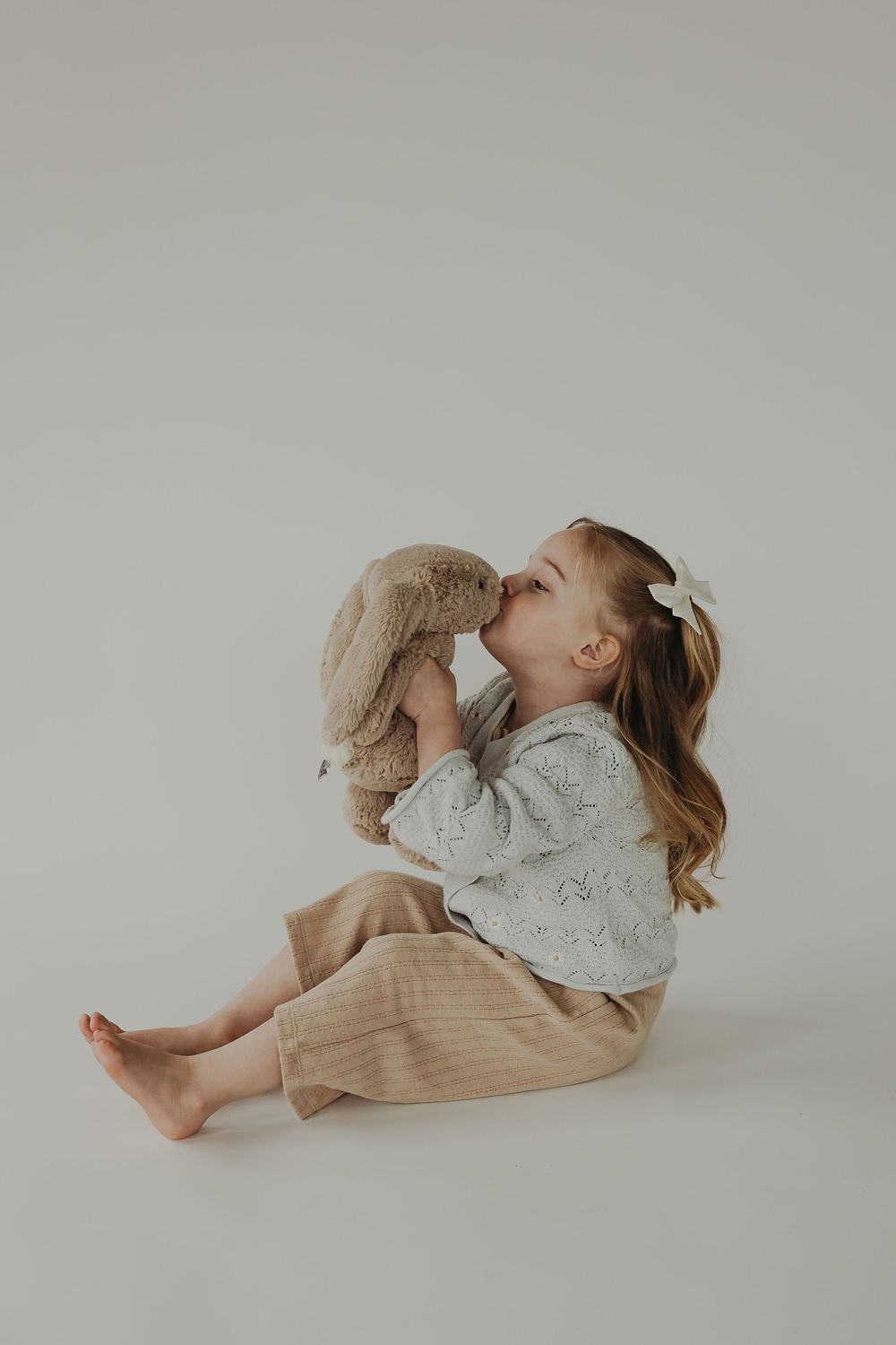 Young child in beige pants and white sweater snuggles with plush bunny toy against neutral background.