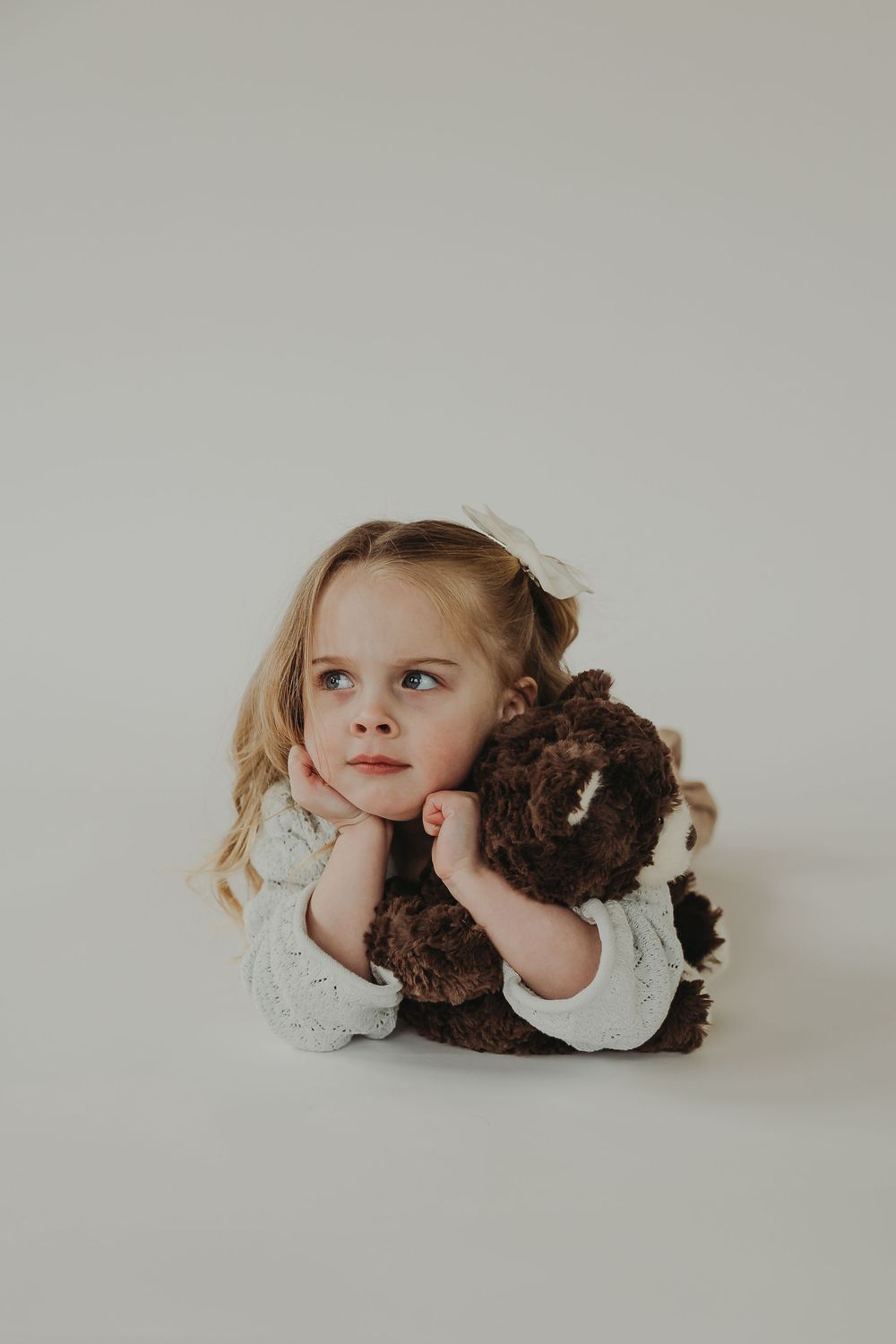 Small child lying on white background with a brown teddy bear showing candid expressions in a minimalist studio setup.