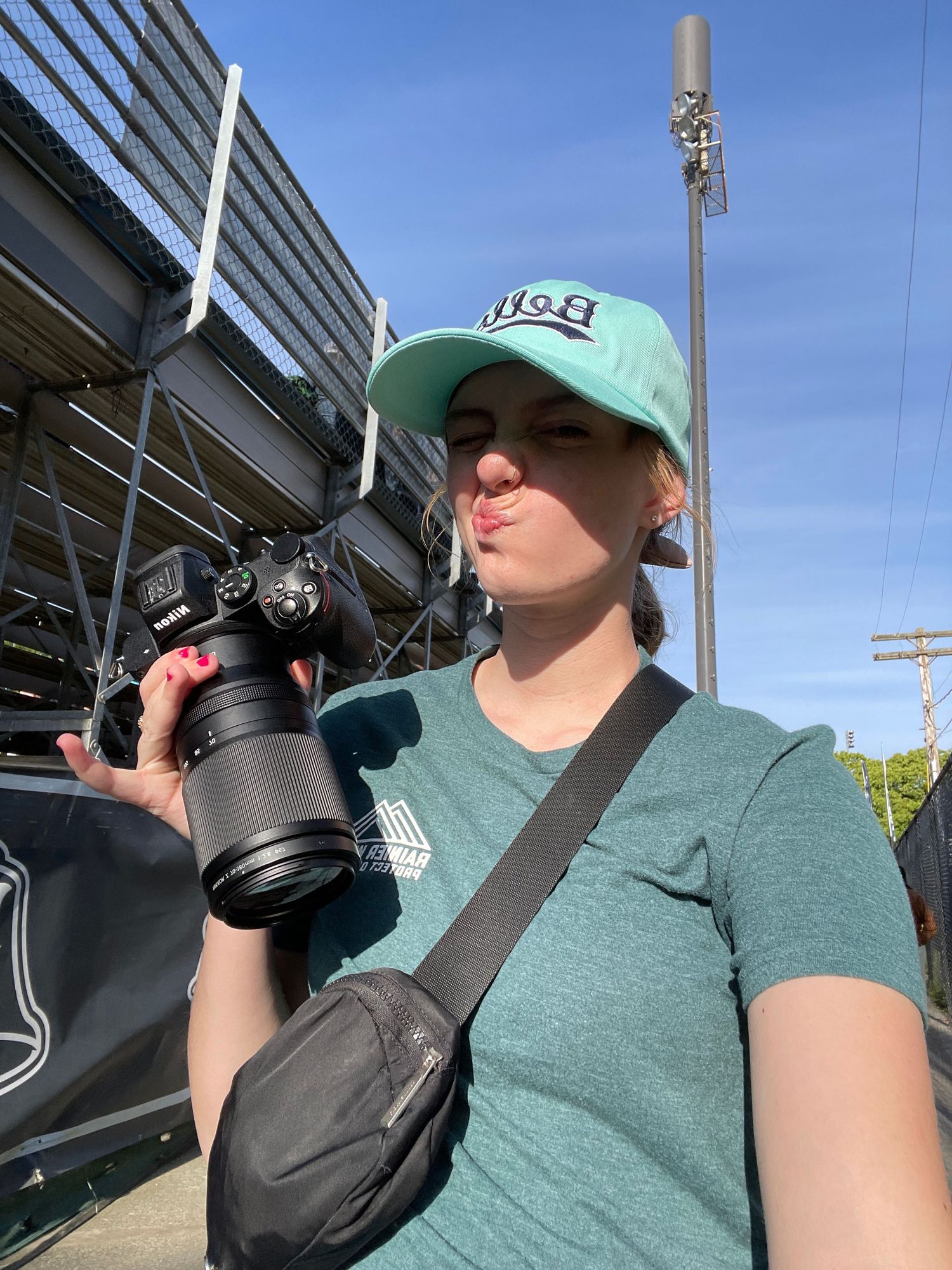 A photographer wearing a green cap takes a selfie with professional camera equipment near a stadium.