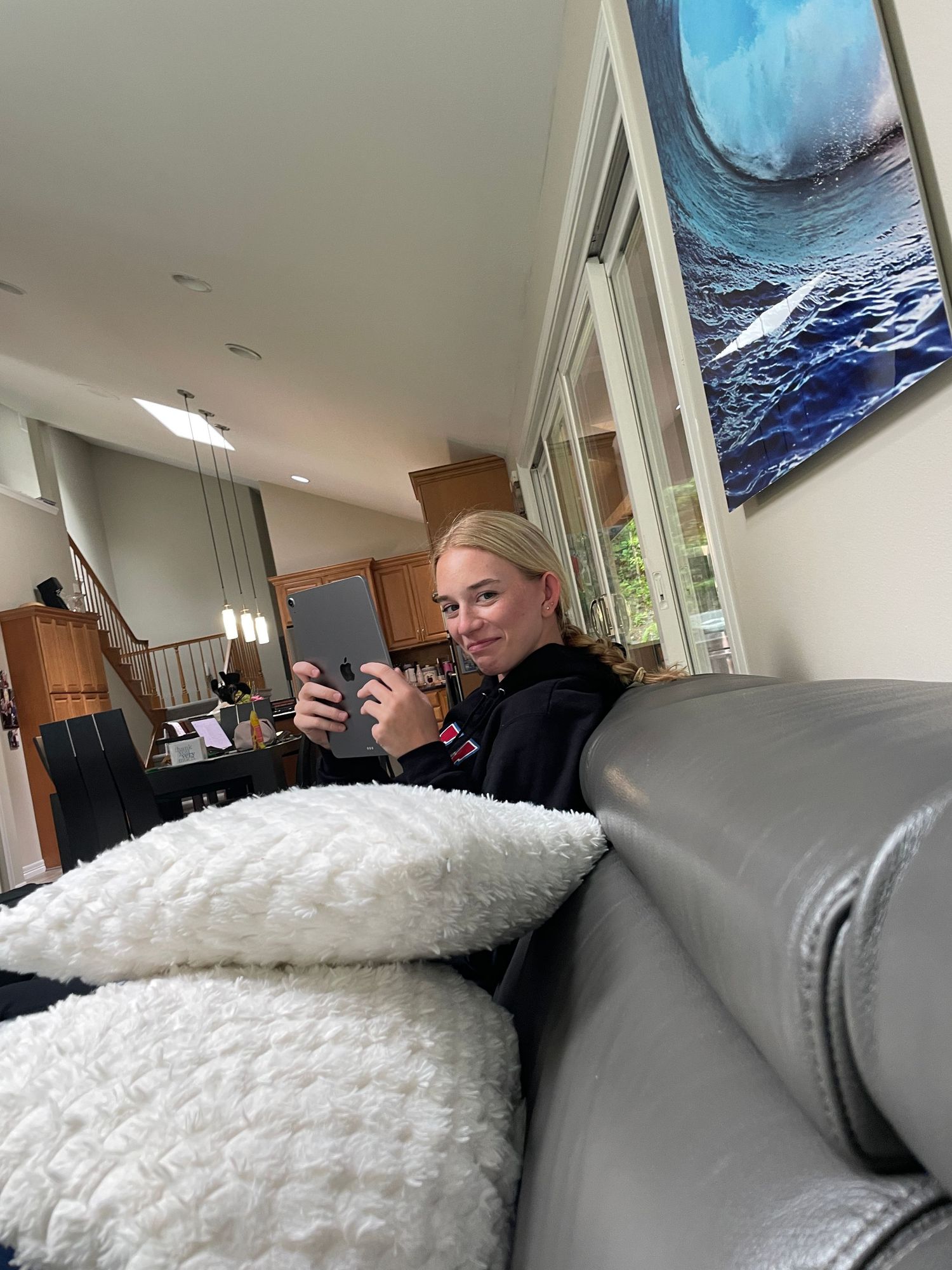 Someone relaxes on a gray couch with a phone, beneath a large ocean wave artwork.