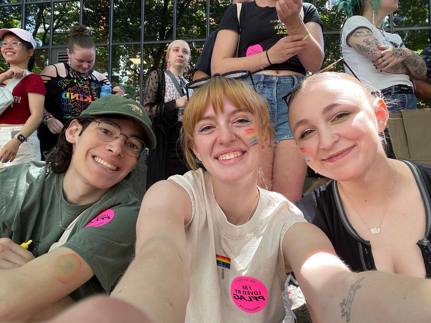 A group of friends taking a selfie together at an outdoor event with pride flags visible.