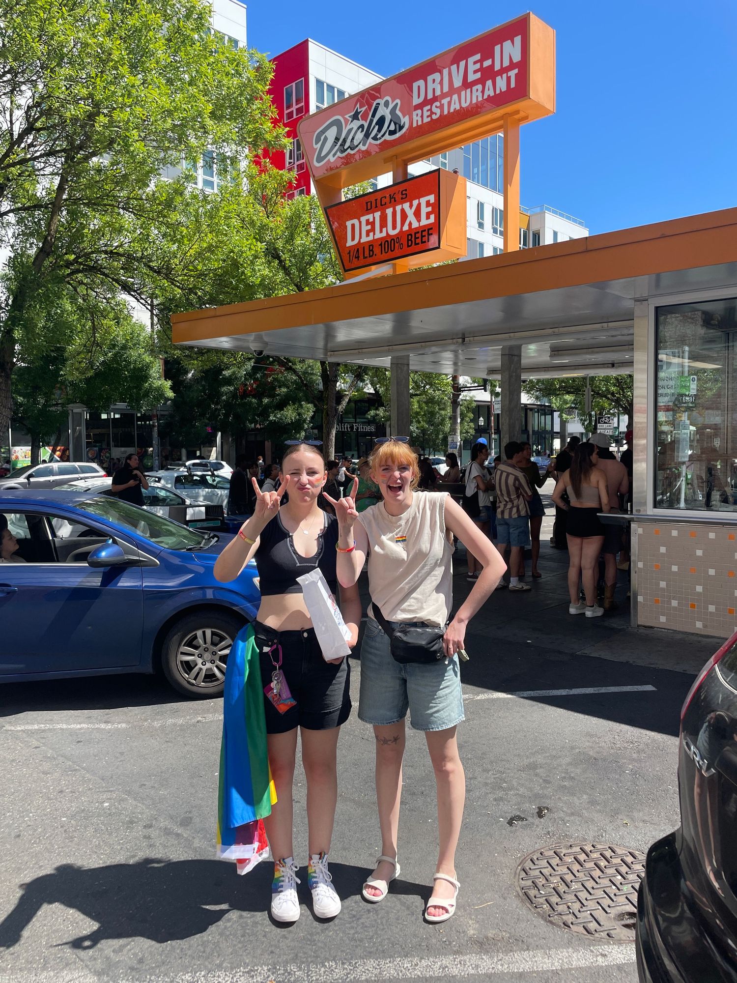 Two people pose together outside a retro-style drive-in restaurant on a sunny day.