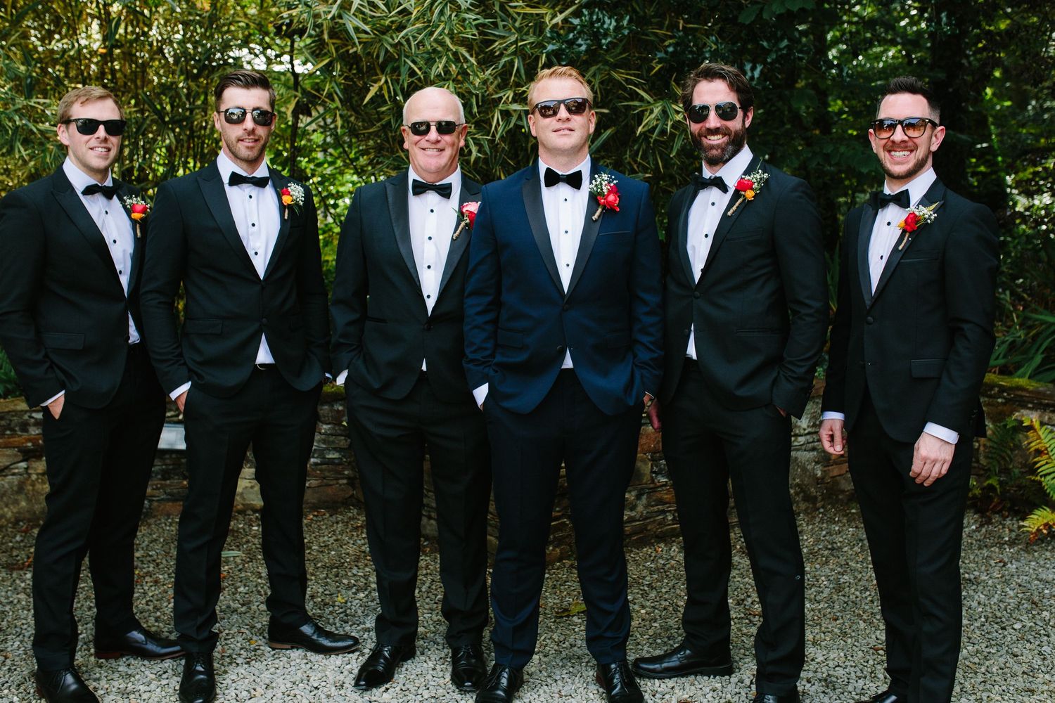 A group of groomsmen wearing black suits with sunglasses posing together outdoors at a wedding.