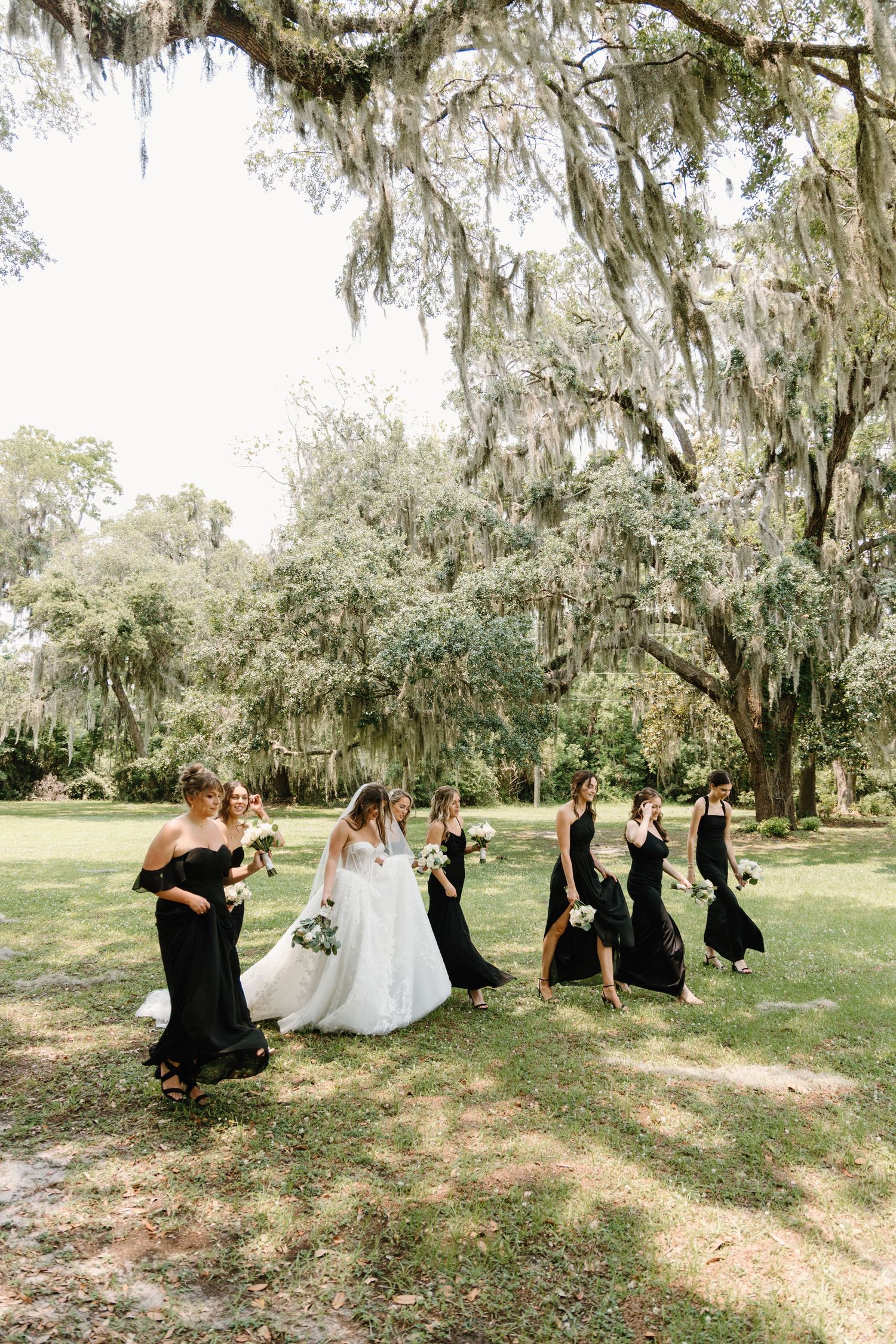 Bridal party in black dresses and white gown poses playfully under Spanish moss draped trees in outdoor photo series.