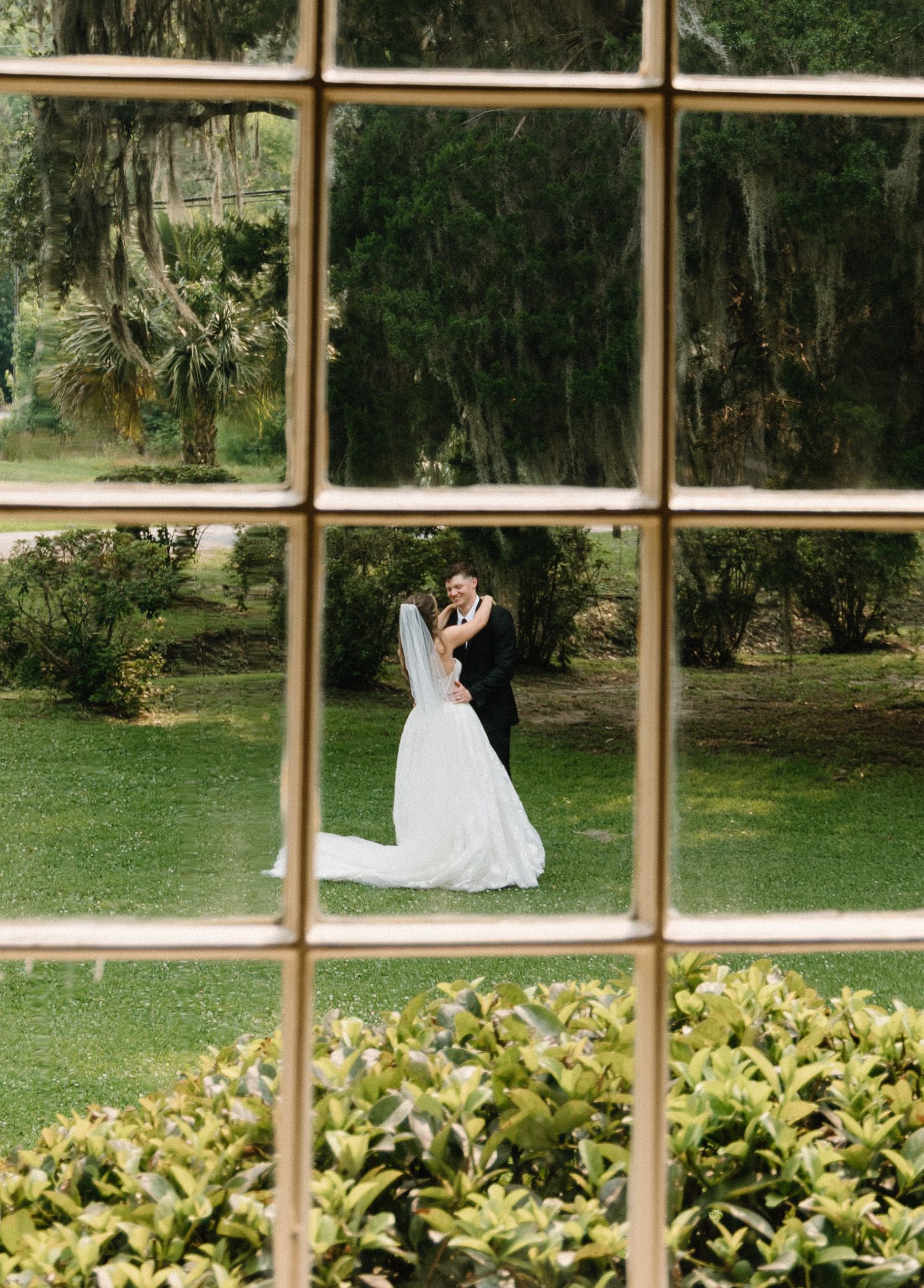 A graceful figure in a white wedding dress twirls on a lush green lawn framed by window panes and garden foliage.