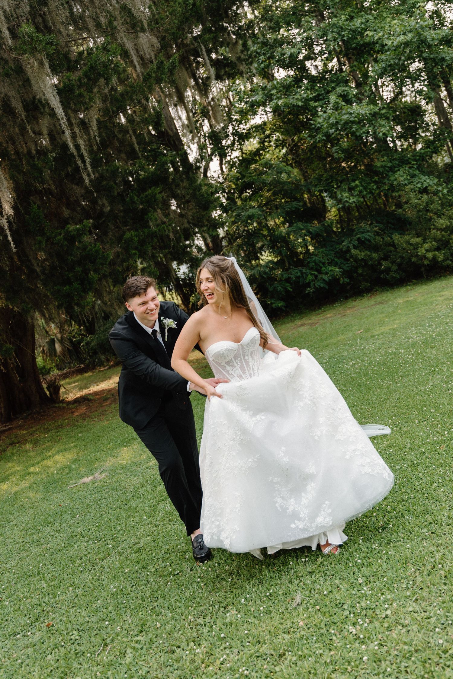 Romantic wedding couple shares playful moment on lush green lawn as bride twirls in white lace gown.
