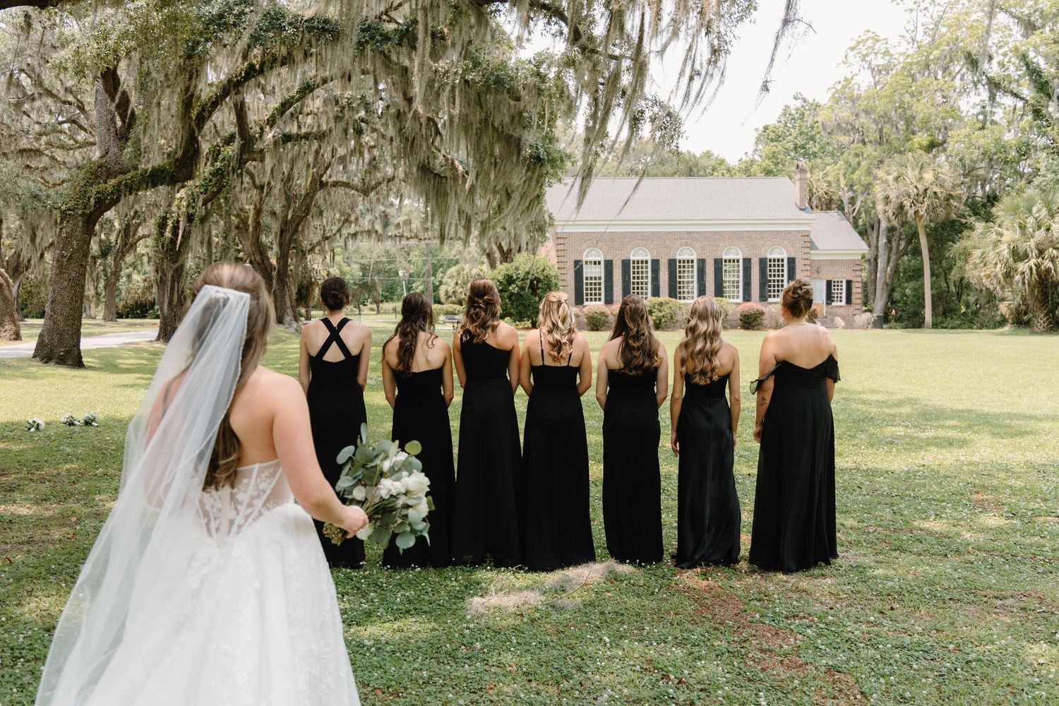 Bridesmaids in long black dresses standing on a lawn with Spanish moss draped trees and a white chapel in background.