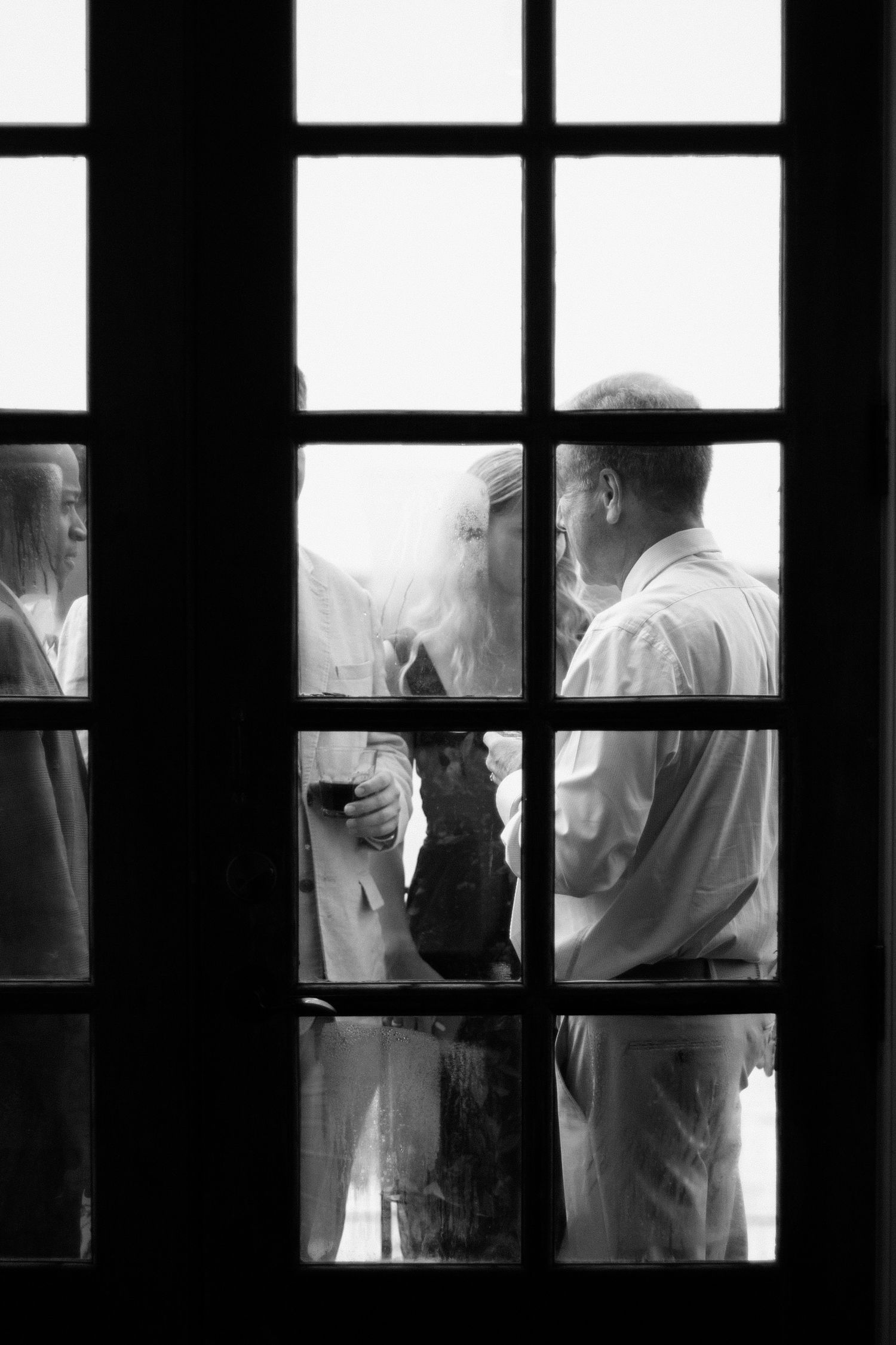 A black and white glimpse through a window shows people gathered in conversation, framed by dark window panes.