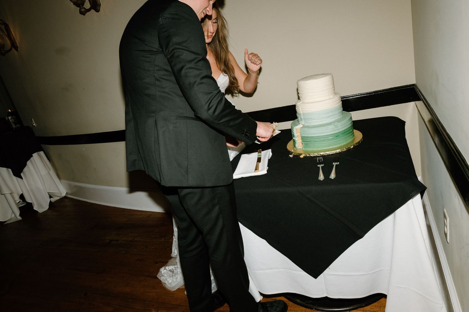 Someone in a dark suit cuts into a three-tiered turquoise and white wedding cake at a reception.