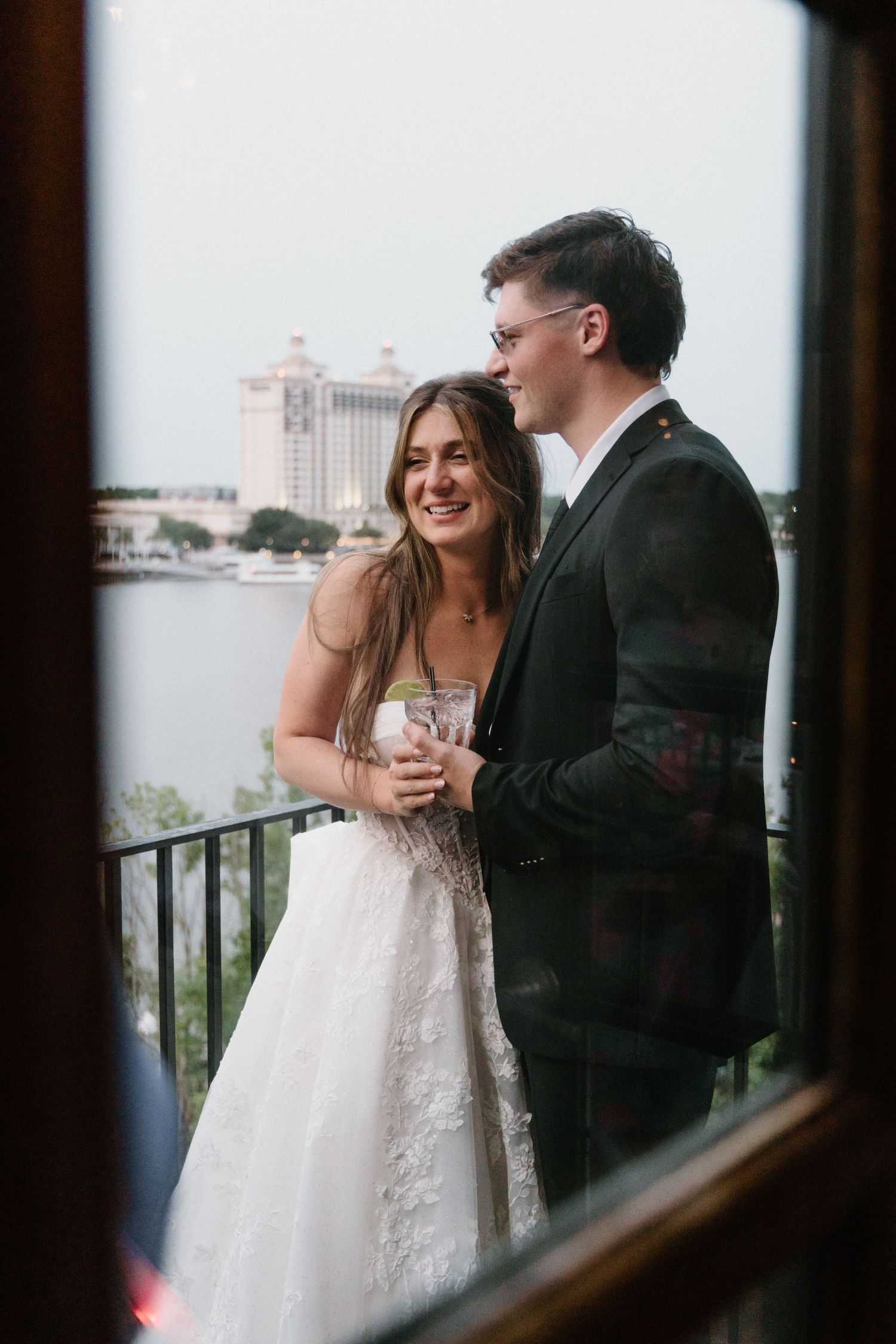 Newlywed couple shares romantic moment on balcony overlooking waterfront with city buildings in background.