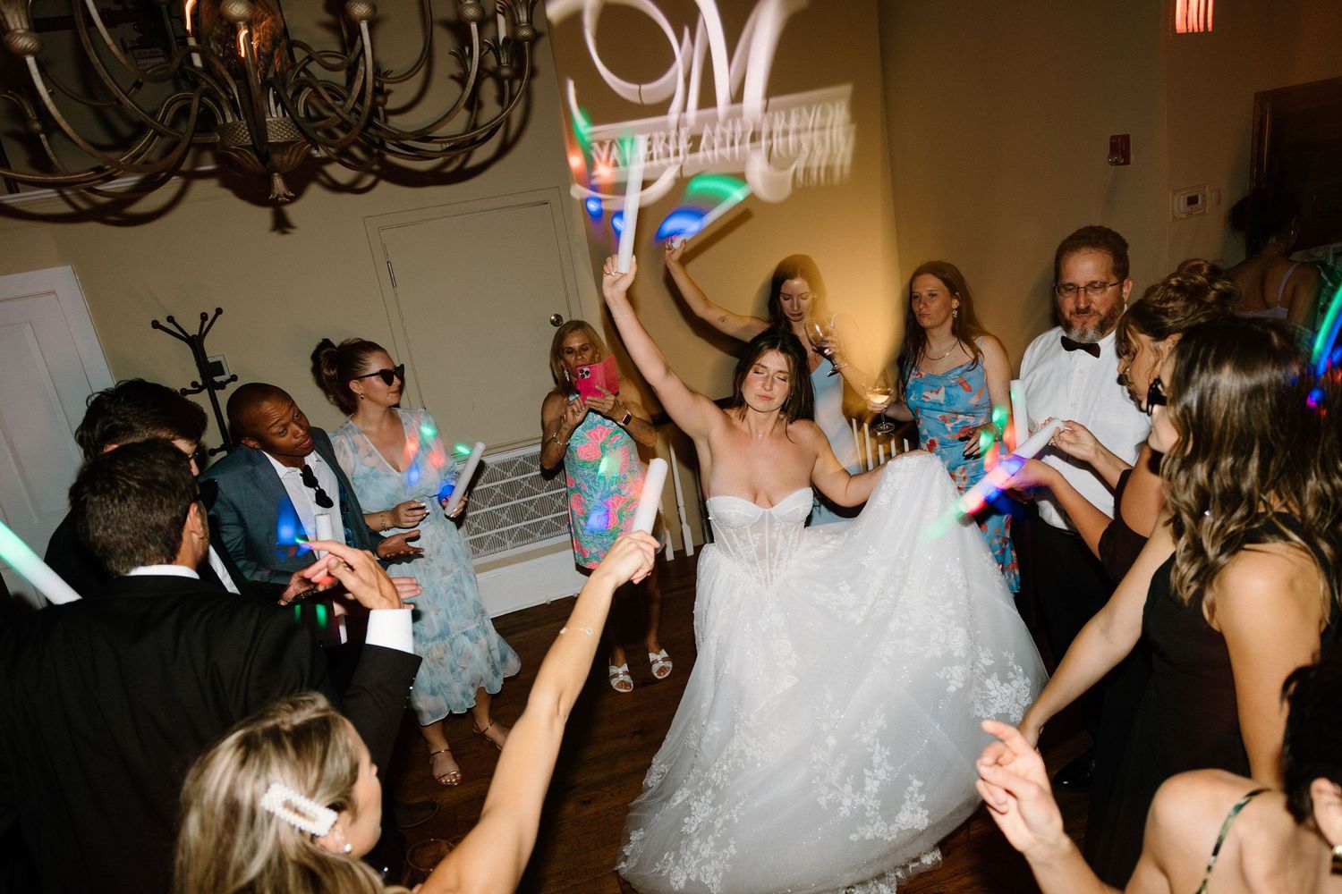 Wedding reception guests dancing and celebrating on the dance floor with glow sticks under a chandelier.