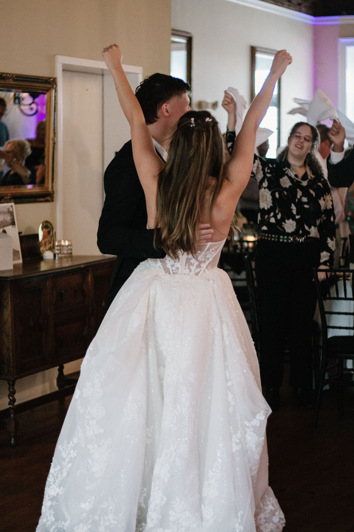 A group of wedding guests dance with their arms raised on a dimly lit dance floor at a reception celebration.