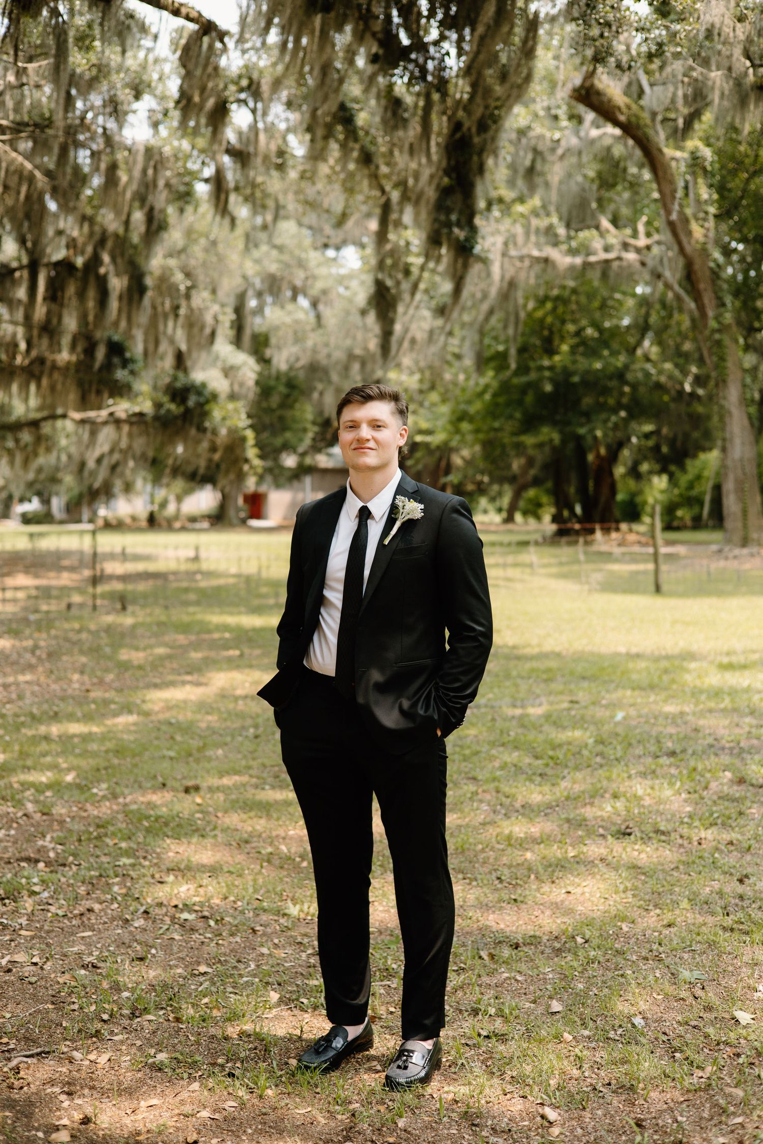 Figure in formal black suit stands in scenic outdoor setting with Spanish moss draped trees and grass in background.