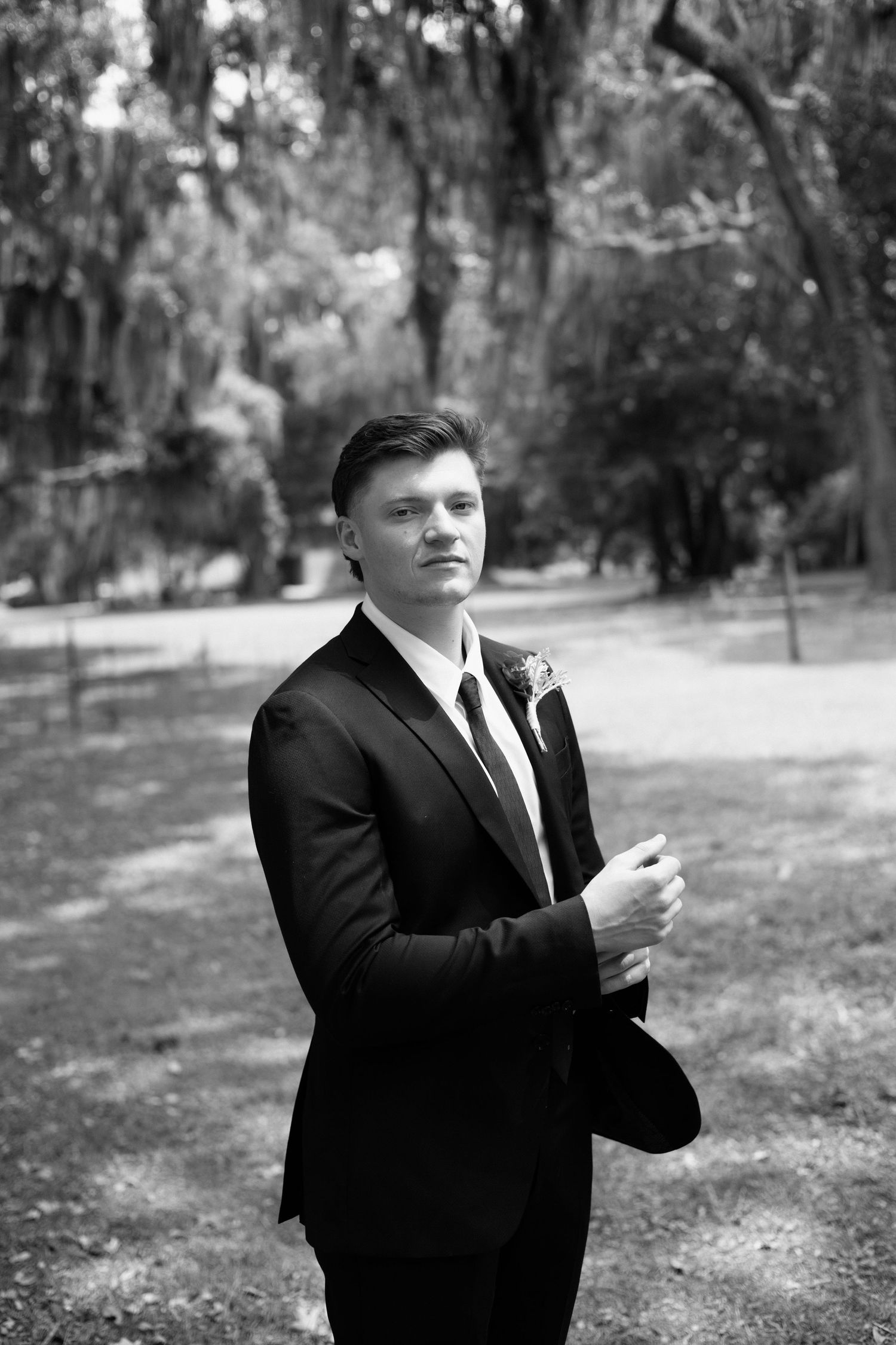 Black and white portrait of a person in a formal black suit and tie standing outdoors among trees in a park setting.