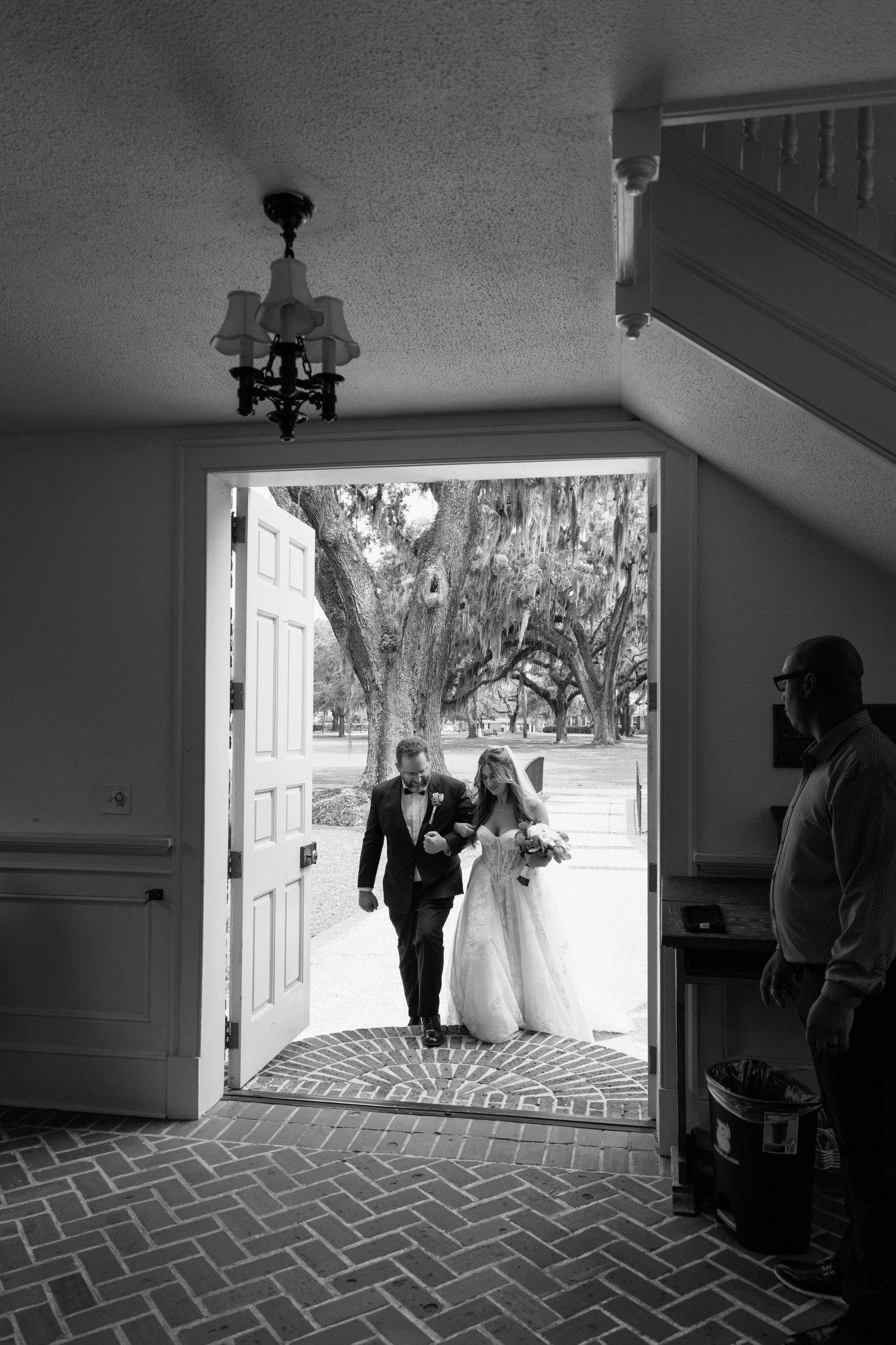 Black and white photo of a couple walking through an elegant doorway with herringbone brick floors and spanish moss in background.