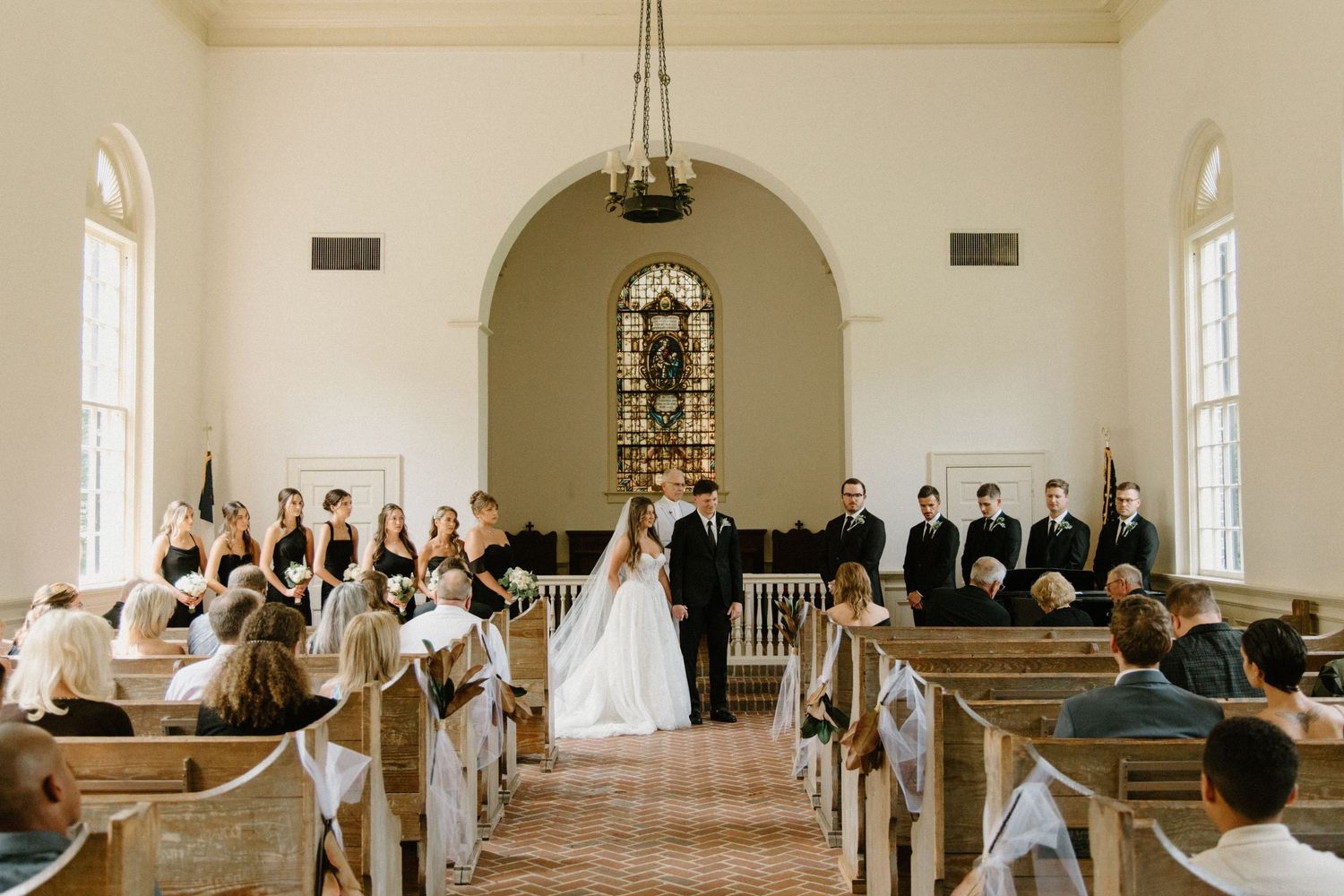A traditional wedding ceremony takes place in a bright white chapel with wooden pews and a stained glass window.