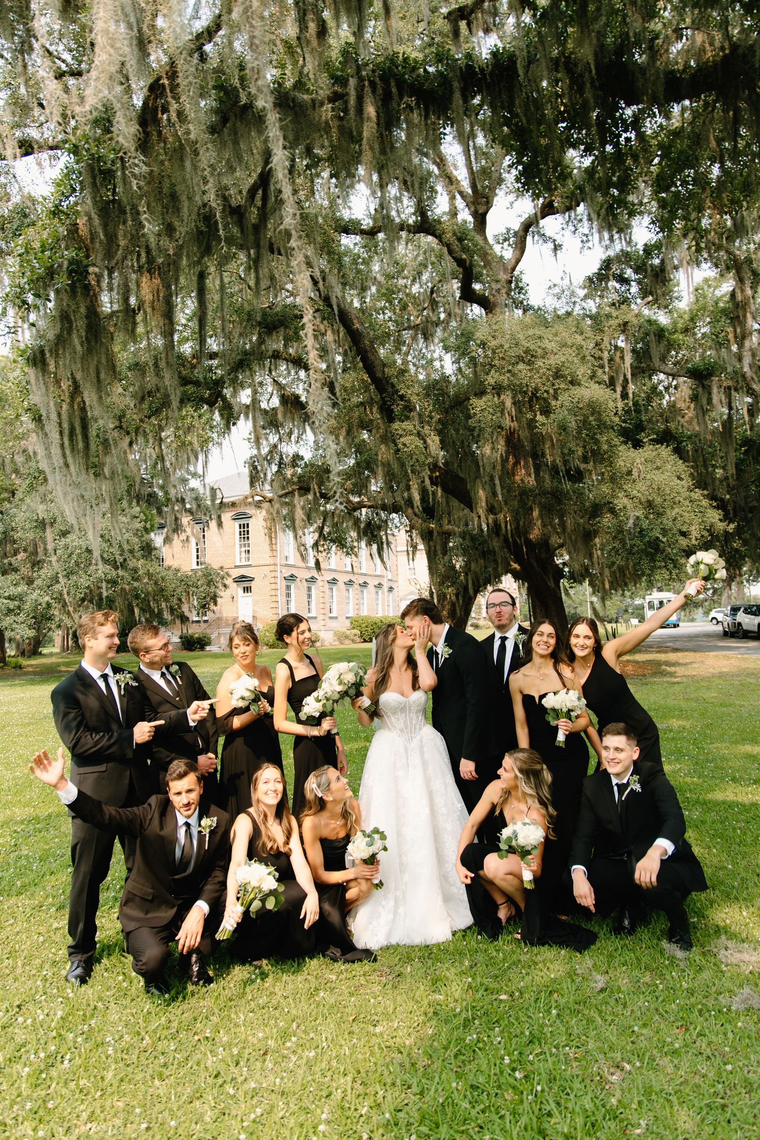 A wedding party sits casually on grass beneath a moss-draped oak tree for an informal outdoor portrait series.