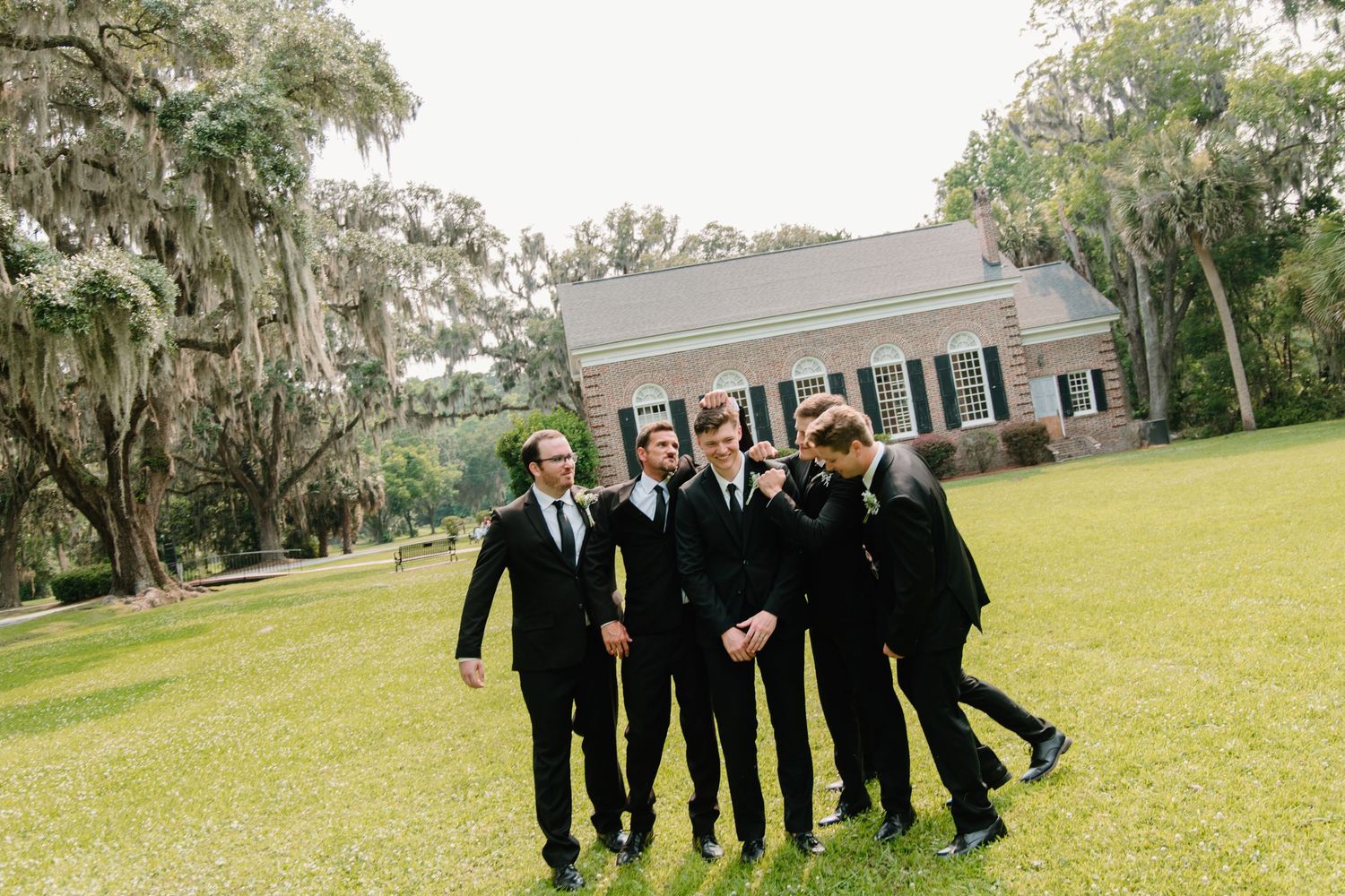 Group in formal black attire pose together on a green lawn with Spanish moss-draped trees in the background.