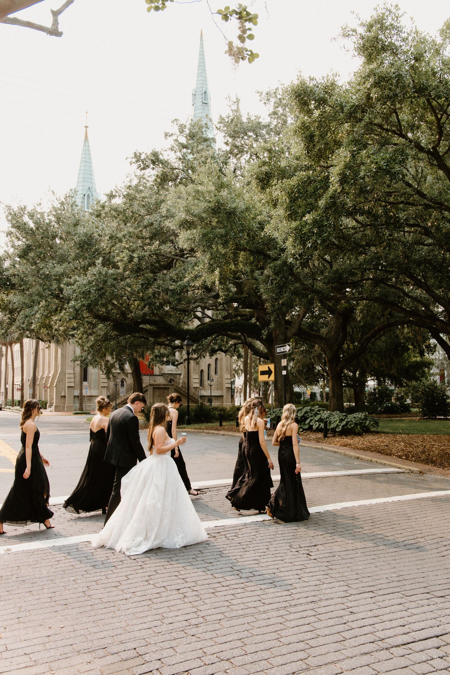 A wedding party in black bridesmaid dresses and white bridal gown poses progressively across brick pathway outdoors.