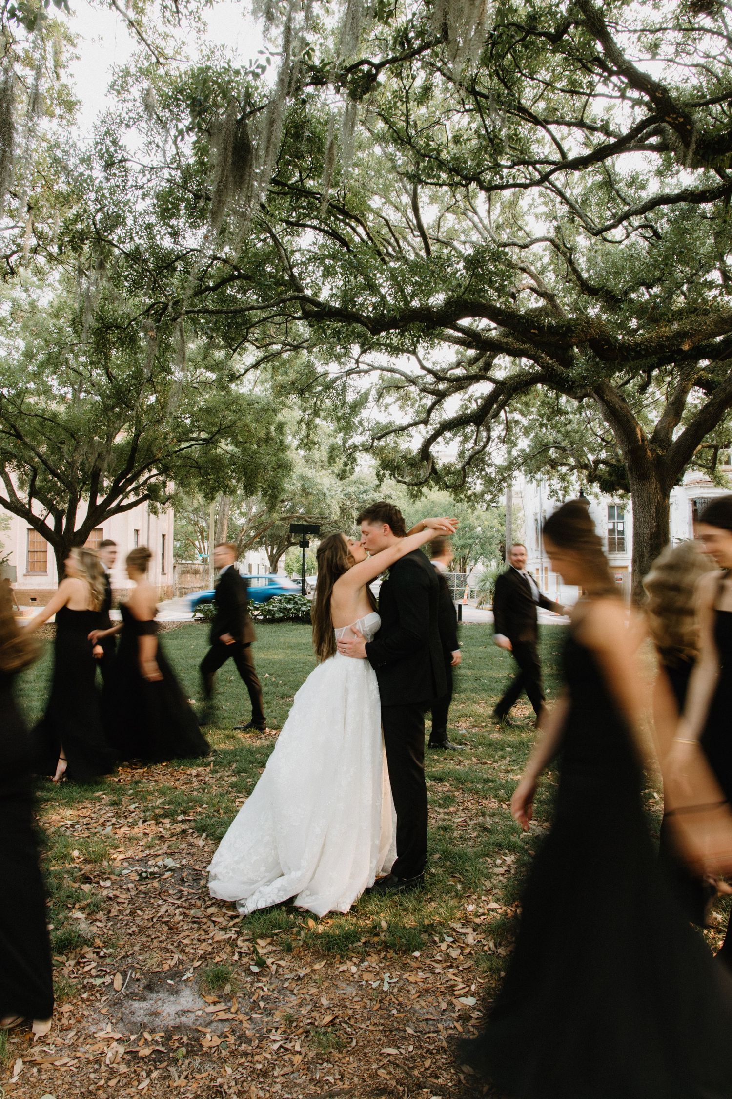 A romantic wedding moment under Spanish moss-draped oak trees as attendants in black dresses surround the dancing couple.