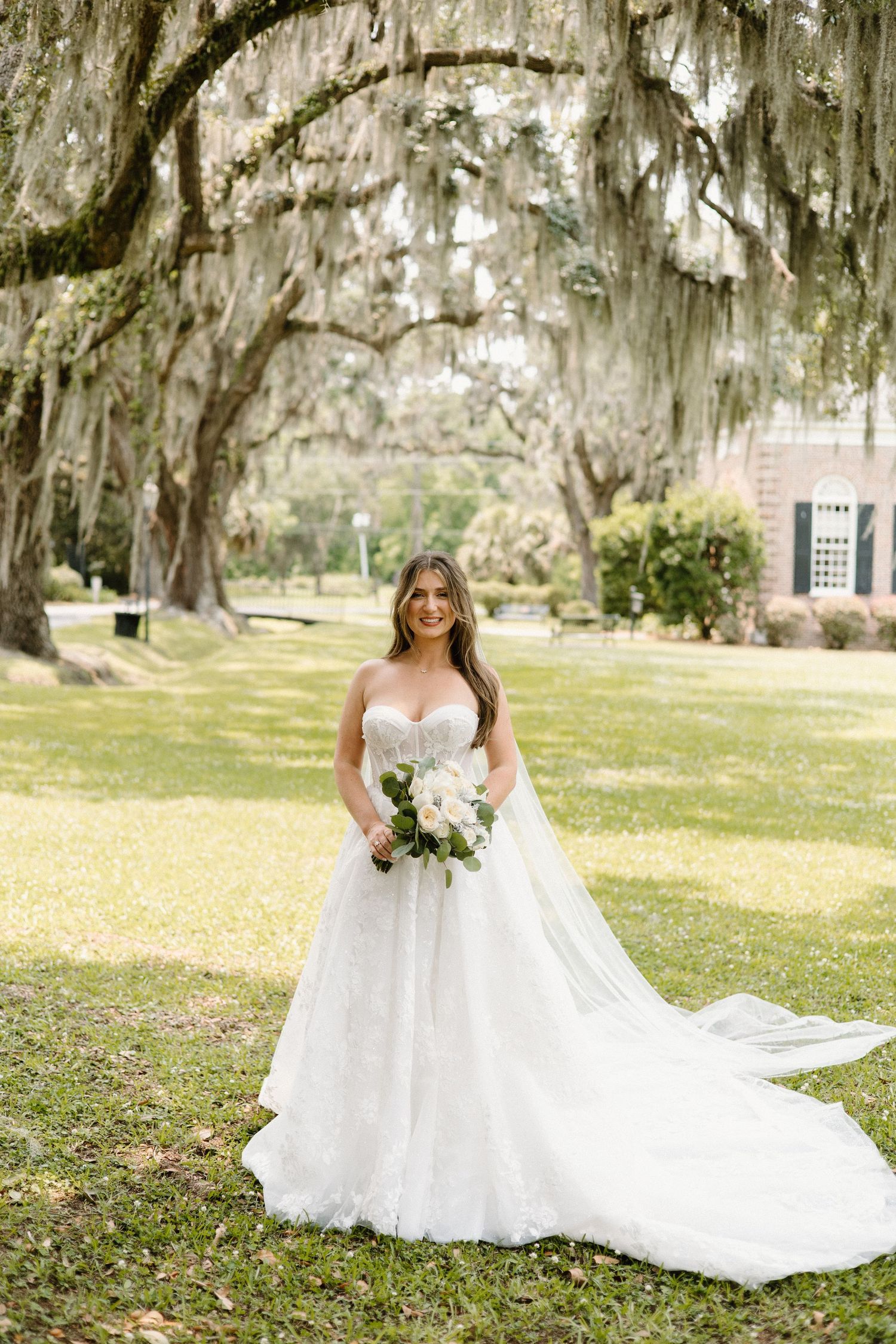 White wedding gown with long train photographed in a scenic garden setting with Spanish moss draped oak trees.