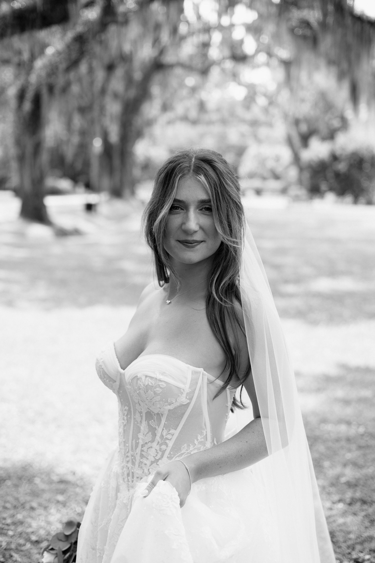 Black and white portrait of a bride in a strapless lace corset dress with flowing veil outdoors in a natural setting.