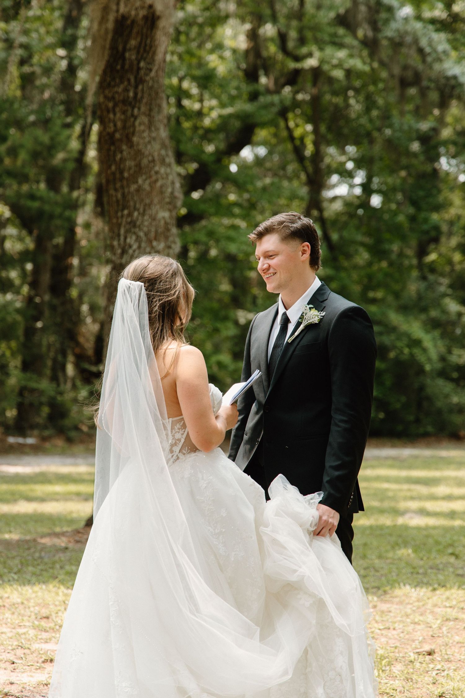 Bride and groom share a joyful moment in a sunlit forest clearing during their outdoor wedding ceremony.