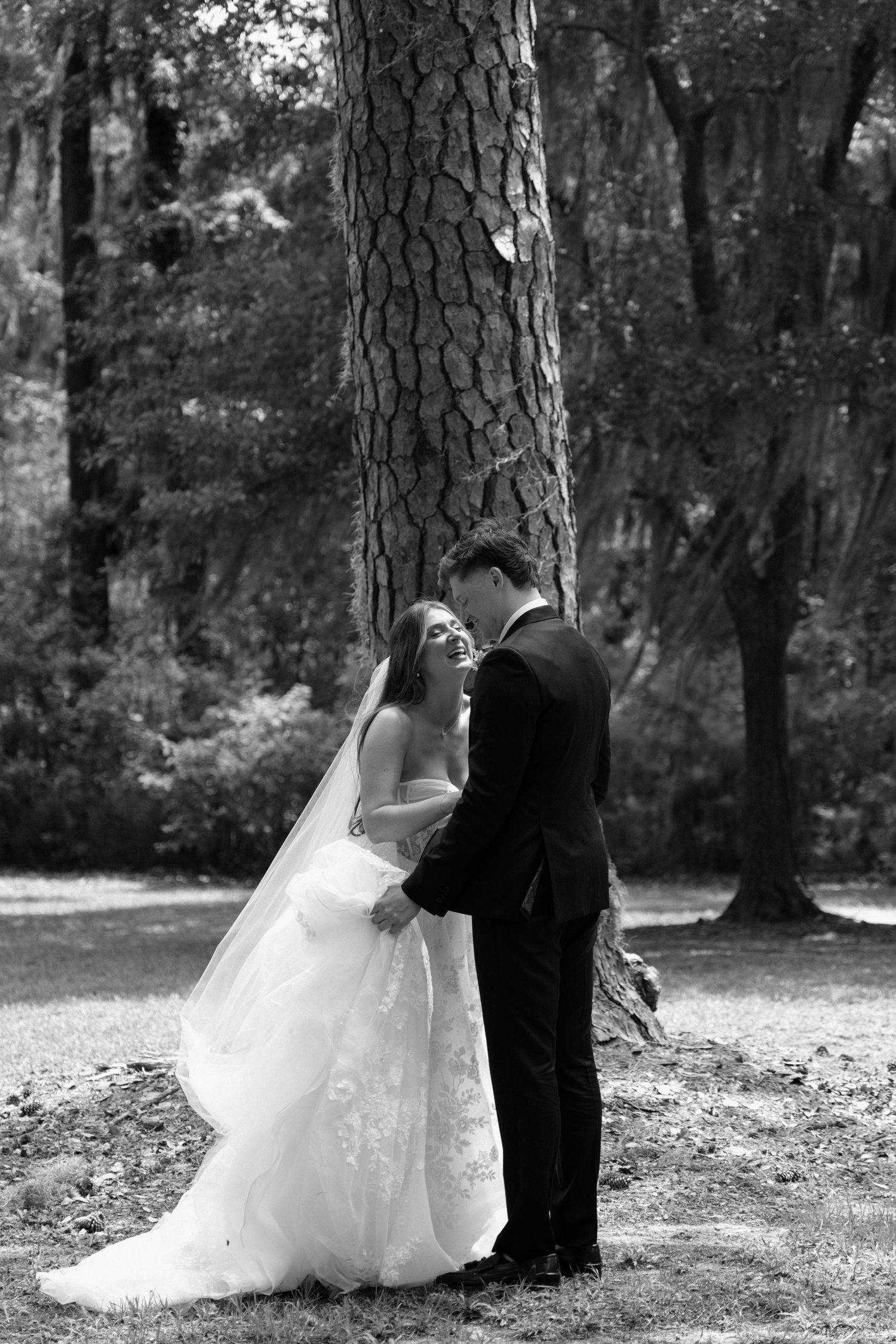 Black and white wedding photo of a couple sharing an intimate moment near a large tree in a forest setting.