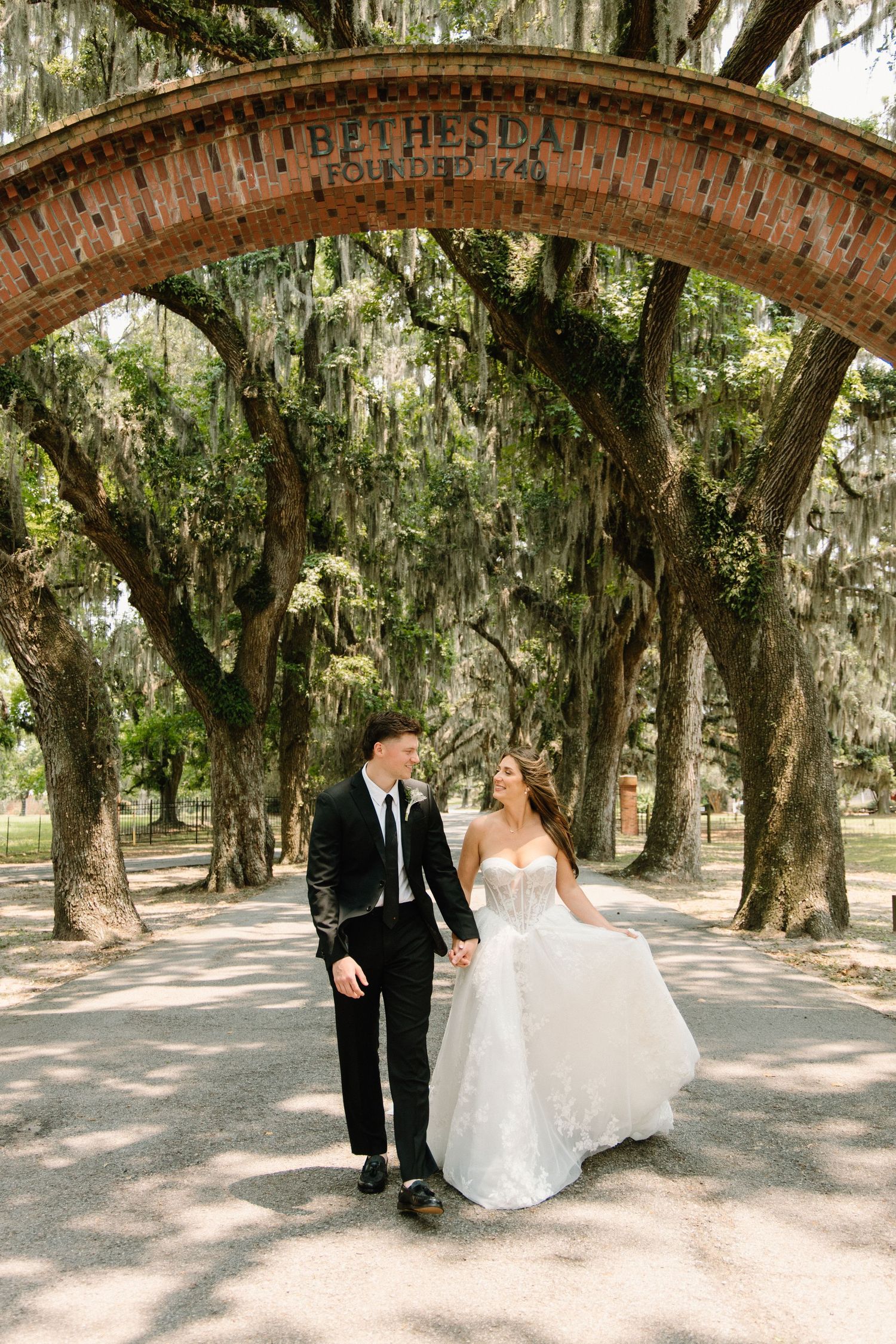 A couple walking under a canopy of Spanish moss-draped oak trees on a path at Wormsloe Historic Site in Savannah, Georgia.