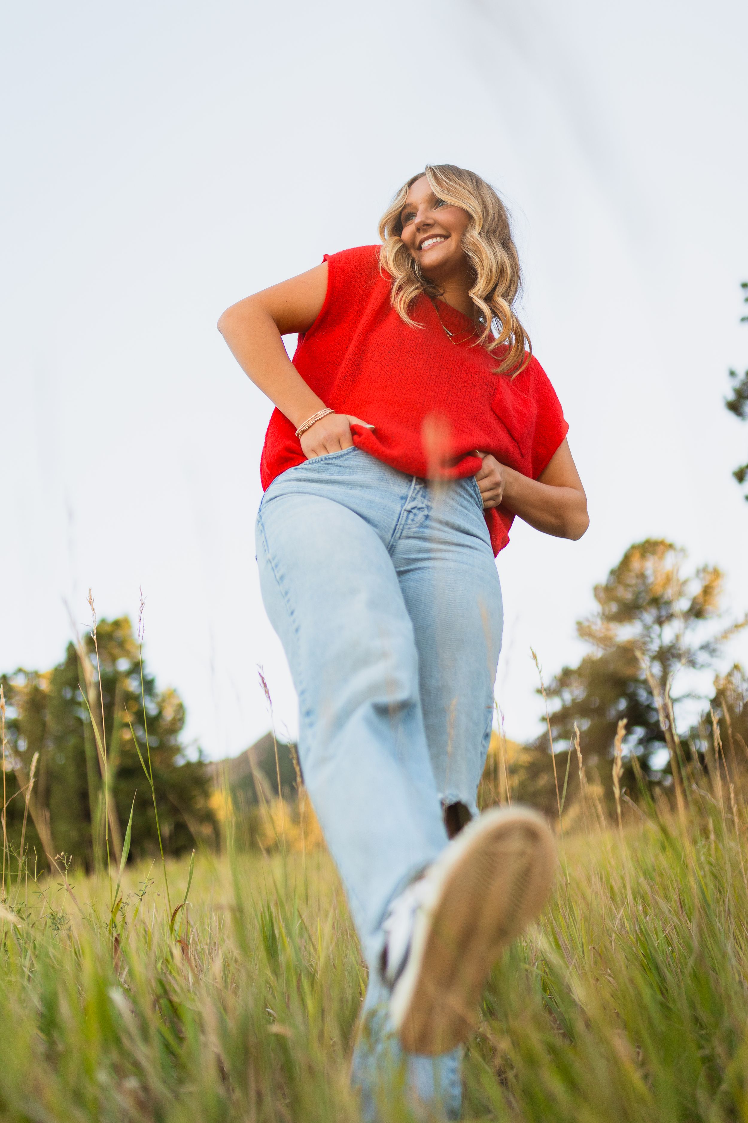 Person in a bright red shirt and light denim jeans stands playfully in a grassy meadow on a sunny day.